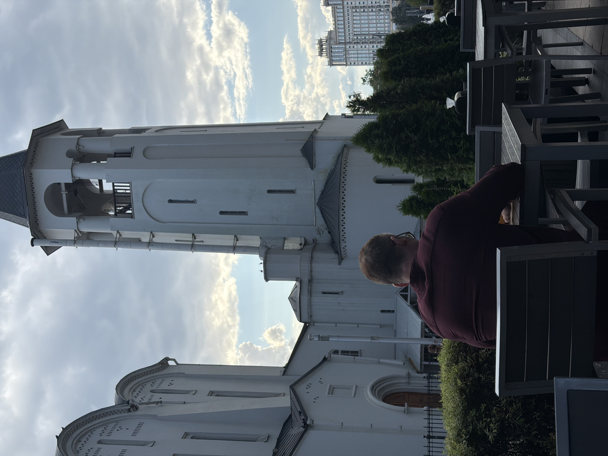 The image shows a man sitting at a table outdoors, facing away from the camera, in front of a large, white building with architectural features reminiscent of a church or bell tower. The building has arched windows and decorative elements. Behind the building, a modern, multi-story building with a patterned facade is visible in the distance. Evergreen trees are planted in front of the white building. The sky is partly cloudy. The man is wearing a dark red shirt and glasses. He appears to be sitting alone at an empty table, with other empty tables and chairs visible in the foreground and background. The scene is likely a public space, possibly a cafe or restaurant with outdoor seating, located in Moscow, Russia, given the architectural style and the presence of the modern building in the background.