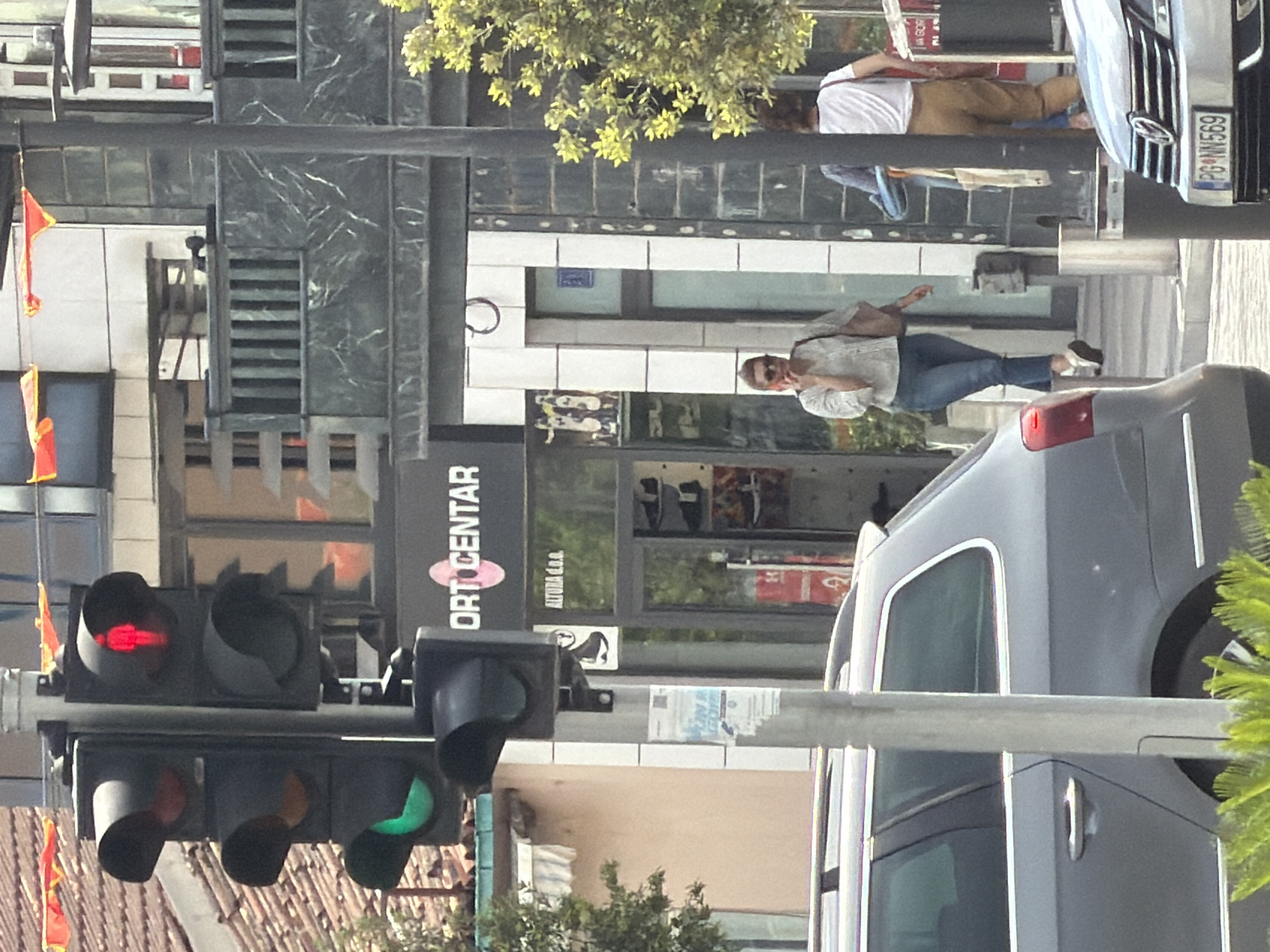 The image depicts a street scene in Podgorica, Montenegro. Traffic lights show green, indicating a go-ahead for traffic. A woman walks on the sidewalk, casually dressed. A pedestrian wearing a white shirt and tan pants stands near a vehicle with a license plate visible; the number is "PG-NN-569." A car, possibly silver or gray, is also prominent in the scene. A sign for "ORTO CENTAR" is visible on a building.