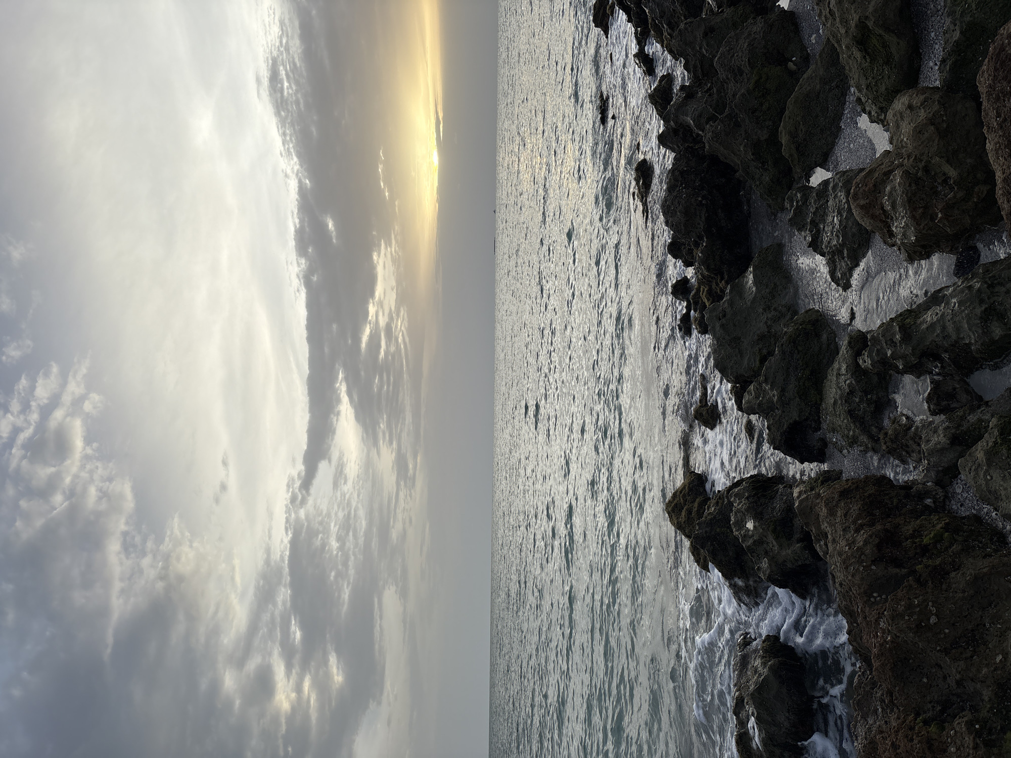 A natural landscape photograph captures a serene ocean scene at sunset. The sky is filled with a mix of dramatic, dark clouds interspersed with softer, lighter clouds, creating a textured canopy. The sun, partially obscured by clouds, casts a warm, golden glow on the horizon, reflecting on the rippling surface of the water. The ocean stretches out to meet the horizon, with small waves gently lapping against a rocky shoreline in the foreground. The rocks are dark and irregular in shape, some covered in a light green moss. The overall impression is one of natural beauty and tranquility, with the elements of sky, sea, and earth harmoniously coexisting. The image does not depict any people, man-made structures, or specific actions. This scene is not located in Venice, United States of America.