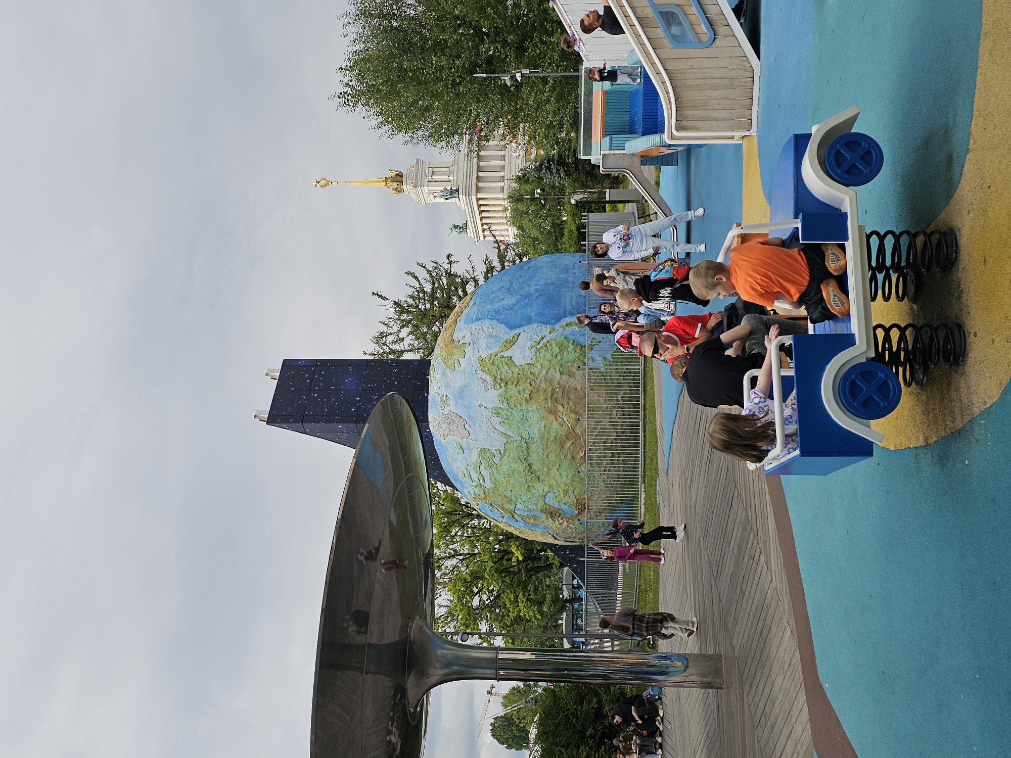 The image captures a lively scene at a playground within VDNKh park in Moscow, Russia. Dominating the background is a large, detailed globe sculpture, with a tall, dark, angular building adorned with a starry sky pattern behind it. To the right, a classic Soviet-era building with a golden spire and white columns stands prominently, its architecture indicative of the park's historical significance.
In the foreground, children and adults are engaged in various activities. A blue toy car, mounted on springs, is occupied by two children. One boy, wearing an orange t-shirt and black shorts, sits in the driver's seat, while a girl in a patterned shirt is seated behind him. The playground itself is colorful, with bright blue and yellow surfaces. Other children are seen playing on a slide and climbing structure to the right. Adults are scattered throughout the scene, some supervising, others observing. The overall atmosphere suggests a bright, cloudy day, with lush green trees surrounding the park area. The wooden boardwalk in the mid-ground adds a natural texture to the urban playground setting.