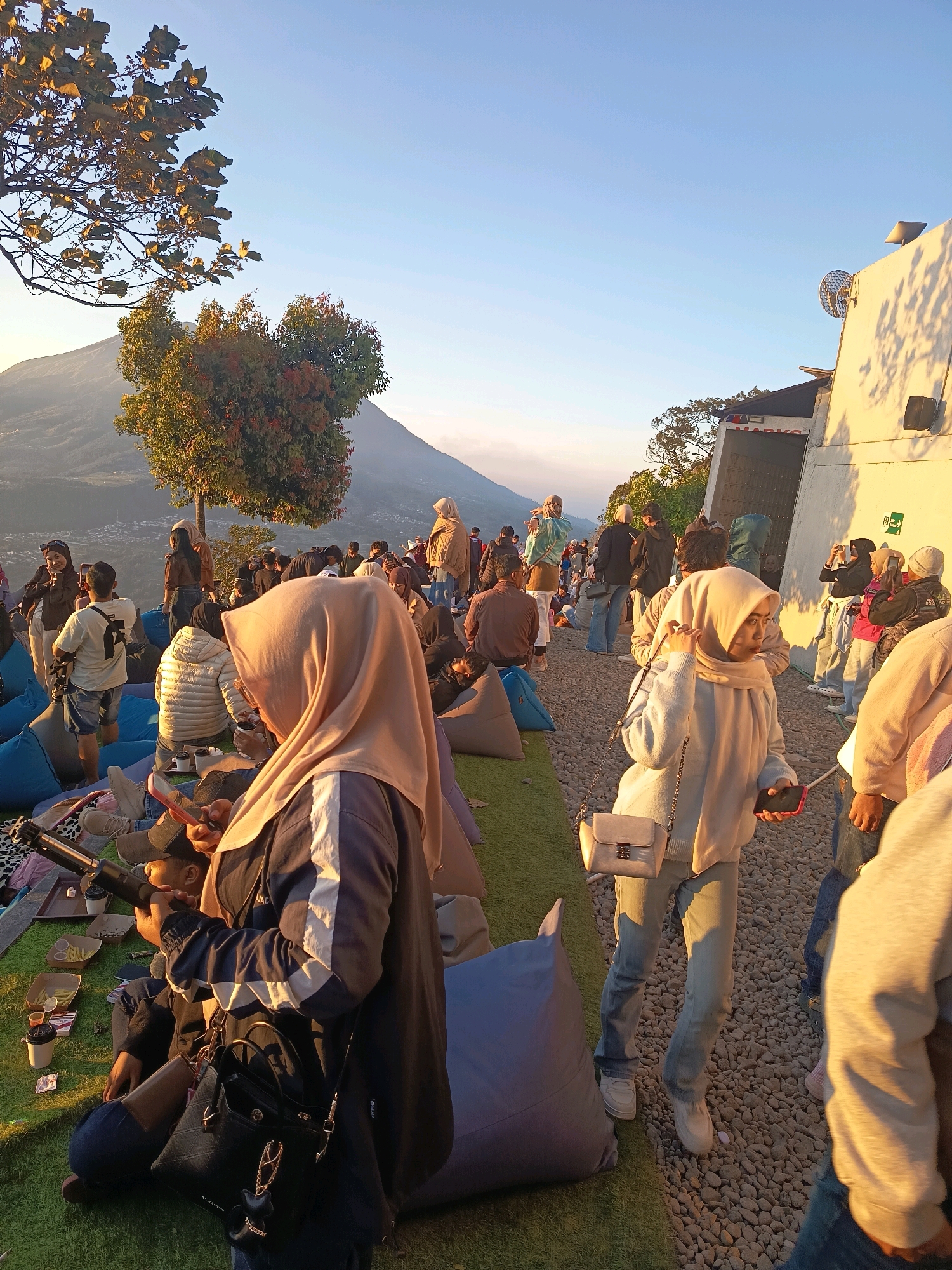This is a photograph of a crowded outdoor gathering on a hillside, likely a tourist spot, at the base of a mountain. The location is identified as Keditan, Indonesia. The time of day appears to be late afternoon or early evening, indicated by the warm, low-angled sunlight casting long shadows.
The scene is bustling with people, many of whom are seated on beanbag chairs and mats placed on a grassy area. The people are dressed in casual clothing, with many wearing headscarves, suggesting a cultural context. Some individuals are holding phones, possibly taking pictures or videos. In the foreground, a person in a dark jacket and a light-colored hijab is seated, holding a phone and what appears to be a monocular or telescope. Another person in the mid-ground, also wearing a hijab, is standing and looking towards the left, holding a phone.
In the background, a majestic mountain rises against a clear blue sky. There are trees on the hillside, some of which are catching the sunlight and appear to have autumn-colored leaves. A building with a white facade is visible on the right side of the frame, with some equipment mounted on its roof and wall, possibly related to broadcasting or observation. The ground is a mixture of grass and small stones. The overall atmosphere is relaxed and social, with people enjoying the scenic view and the company of others.