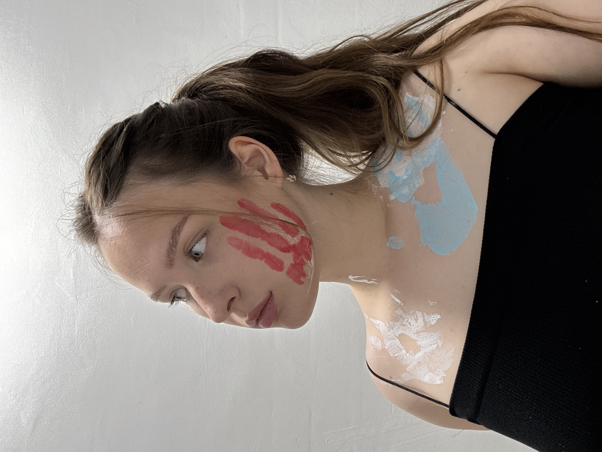 The image depicts a young woman with light skin and brown hair, styled in a ponytail. She is wearing a black spaghetti strap top. Her face and upper body are partially covered in paint. There are red handprints on her cheek and neck, and patches of light blue and white paint on her neck and upper chest. The woman is looking downwards and to her left, with a neutral expression. The background is a plain, textured, light-colored wall, suggesting an indoor setting. There is no visible text. The lighting appears to be from a single source, casting subtle shadows. The image is cropped to focus on the woman's upper body and face, with her hair extending out of the frame. The context of Chaikovsky, Russia, does not provide specific visual cues within the image itself.
