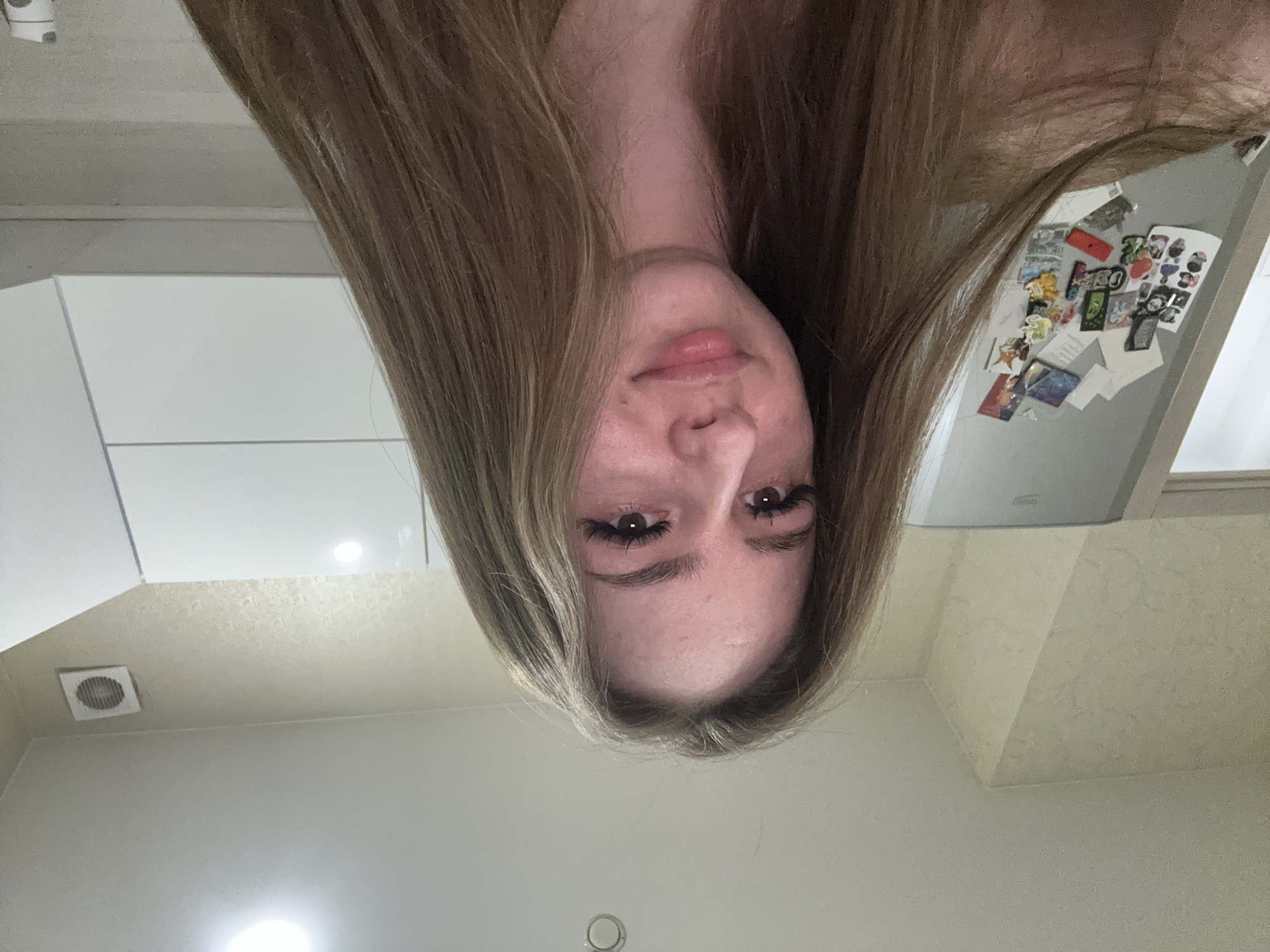 A person with long, light brown hair is upside down, positioned in front of what appears to be a kitchen environment. The person's face is centered, with visible eyes, nose, and lips. In the background, there are white cabinets, a refrigerator covered with magnets, and a white ceiling. The scene's location is Unknown, Unknown.