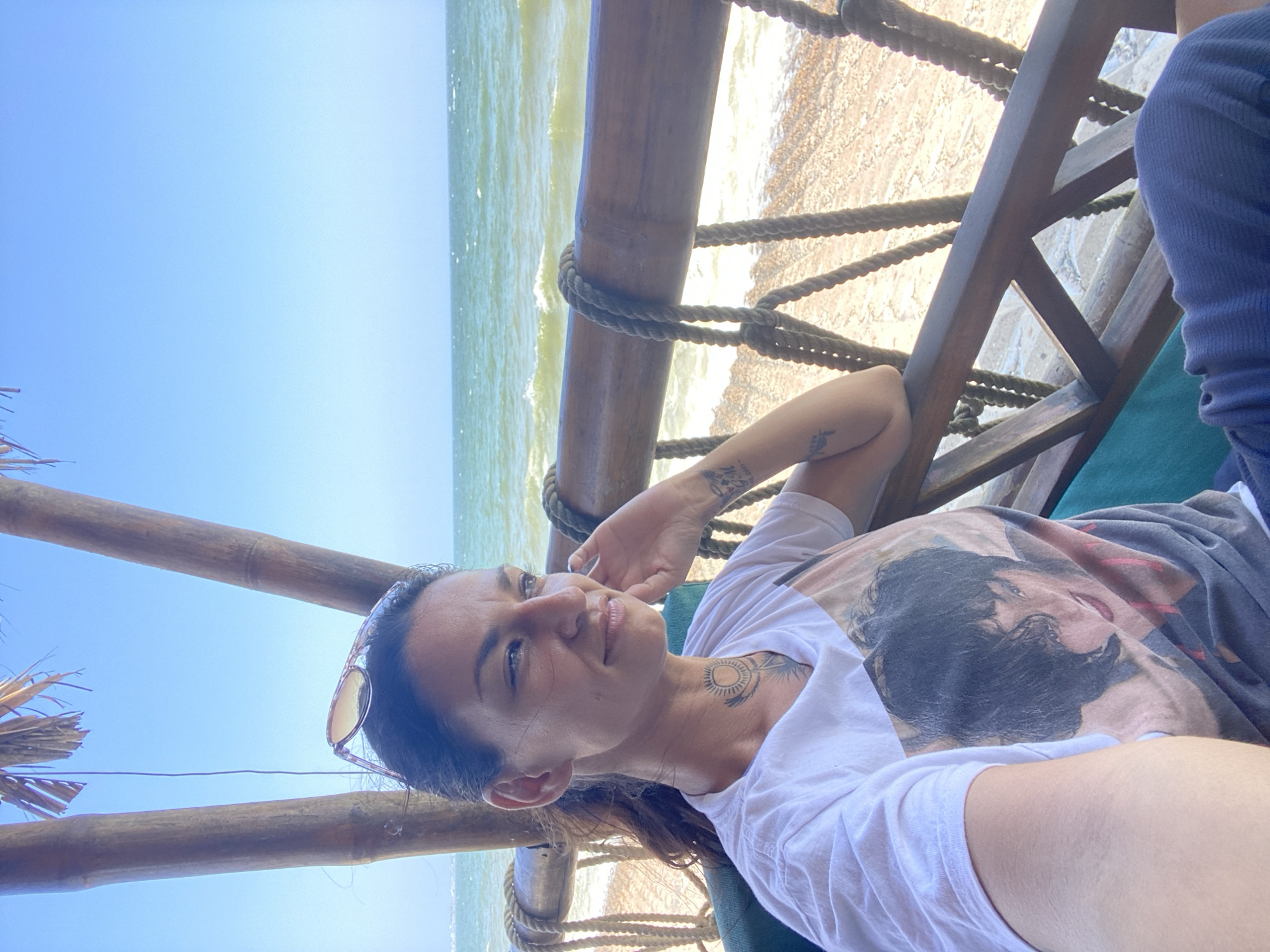 A woman sits on a chair facing a body of water, likely the ocean, in Mui Ne, Vietnam. She is wearing a white t-shirt with a picture of a person on it, sunglasses perched on her head, and dark pants. The scene is set on a beach with a wooden structure and bamboo poles providing shade. The water is a vibrant green with gentle waves, and the sky is a clear blue. The woman is looking away from the camera, with her hand resting on her chin, suggesting a relaxed or contemplative pose. The presence of the beach, ocean, and tropical vegetation points to a vacation or leisure setting.