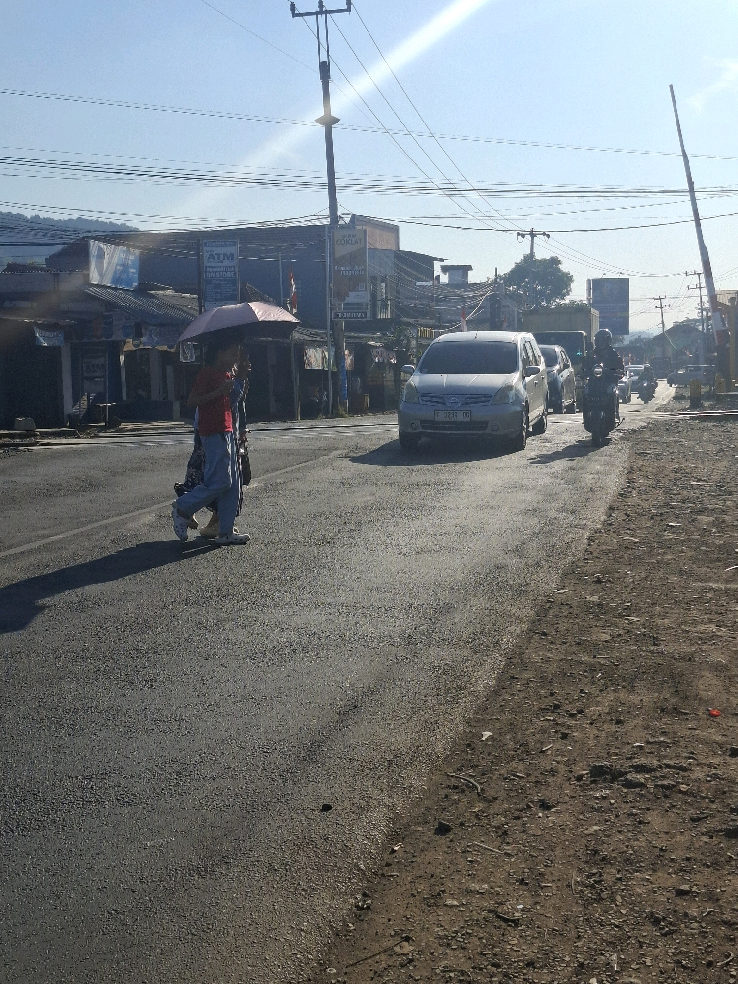 The image depicts a street scene in Lembursawah, Indonesia, under a bright, sunny sky with visible sun flare. In the foreground, a paved road stretches into the distance, bordered by a dirt shoulder. Two individuals are walking on the left side of the road, one holding a pink umbrella. A silver minivan is driving on the road, followed by other vehicles, including a motorcycle with a rider wearing a helmet. Along the left side of the road, buildings with awnings and various signs, including "ATM" and "TOKO MUTIARA", line the street. Overhead, a network of power lines and a utility pole are prominent. A railroad crossing gate is visible on the right side of the image. The overall scene suggests a typical day in a semi-urban or commercial area with ongoing traffic.
