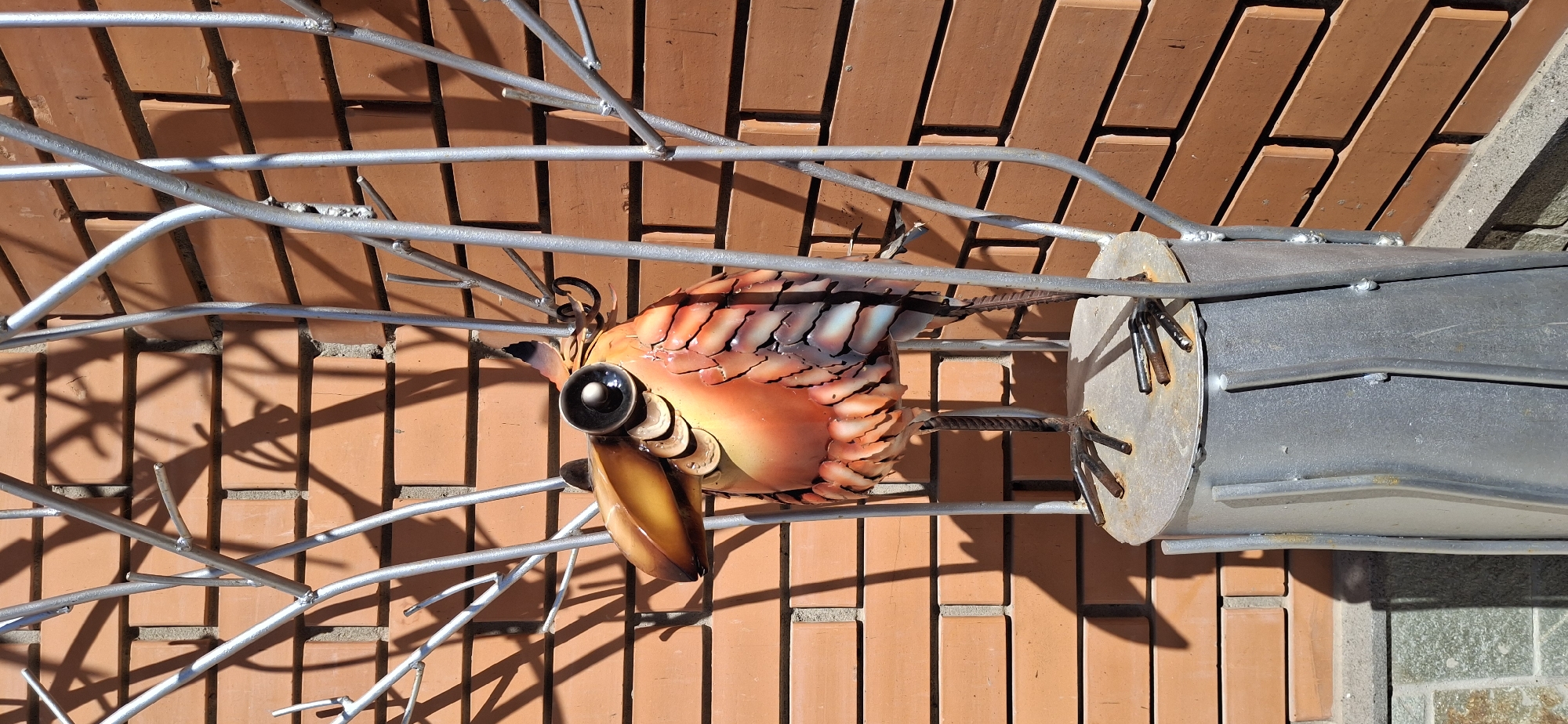 A metal sculpture of a fish, rendered in vibrant orange and yellow hues with light blue accents on the tail scales, is mounted on a metal frame resembling branches. The eye of the fish is a prominent black sphere. The sculpture is positioned against a backdrop of a red brick wall and some grey tiles. The photo was taken in Izhevsk, Russia.