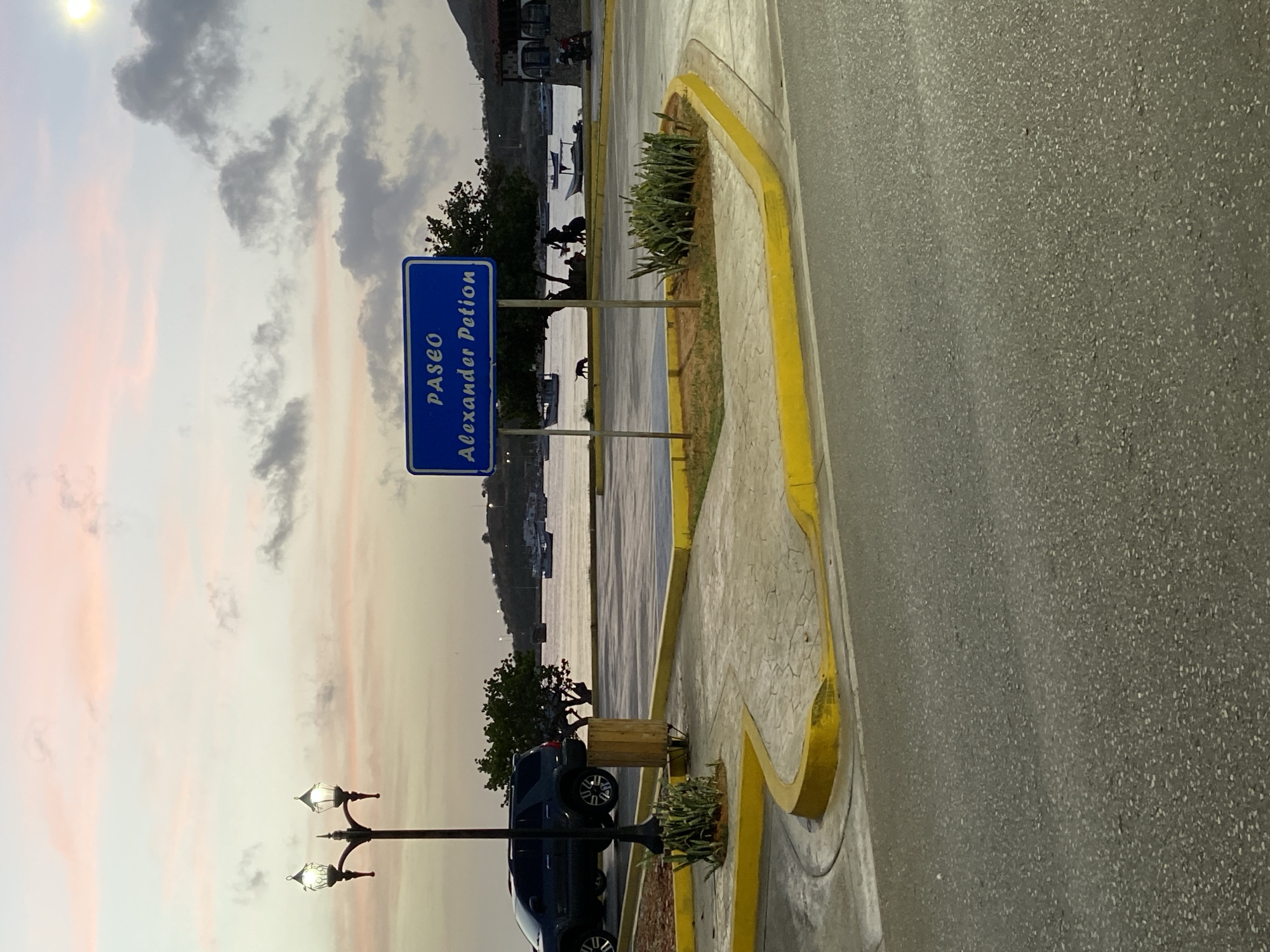 The image shows a street scene in Juan Griego, Venezuela, at sunset. A blue sign with white lettering reads "PASEO Alexander Petion." The sky is a gradient of pink and pale yellow, with scattered grey clouds. In the foreground, there is a paved road and a sidewalk with yellow curbs. A dark-colored SUV is parked on the left side of the frame. A lamppost with two lights stands on the sidewalk. Further back, a body of water, likely the sea, is visible with several boats and a distant shoreline with buildings and hills. A few people are gathered near a tree by the water, and a dog is walking on the beach.