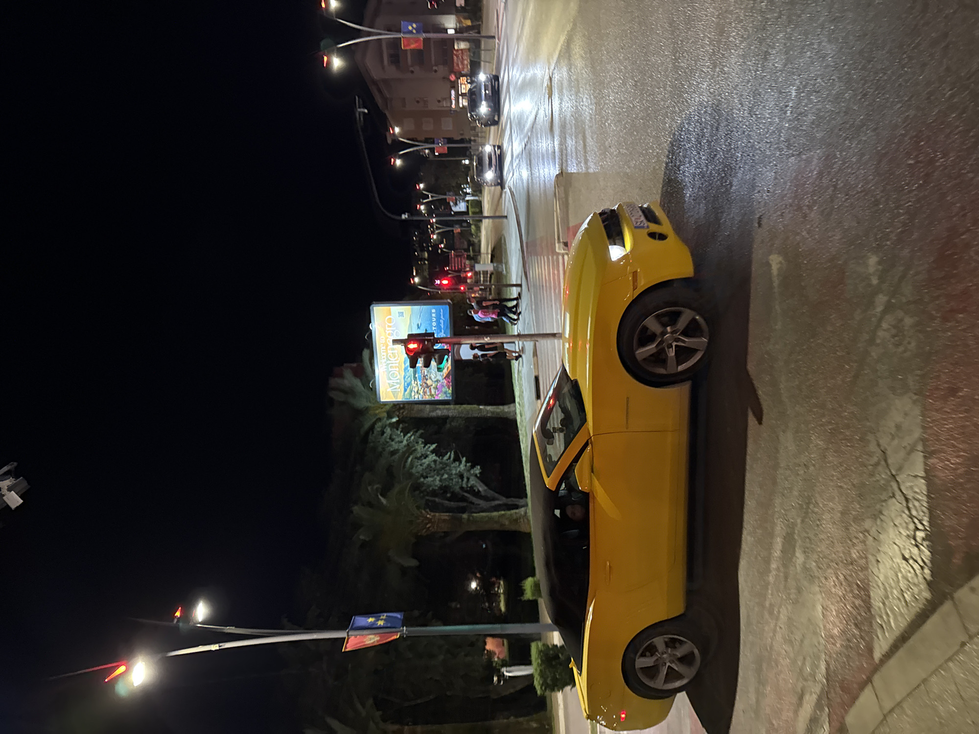 A yellow convertible car sits on a wet road in the city of Budva, Montenegro, at night. The street is lit by streetlights. In the background, there are buildings, pedestrians, palm trees, and an illuminated billboard for Montenegro Tours. Traffic lights are visible, showing red. Further back, other cars are on the road. Flags are attached to a pole, partly obscured by foliage.
