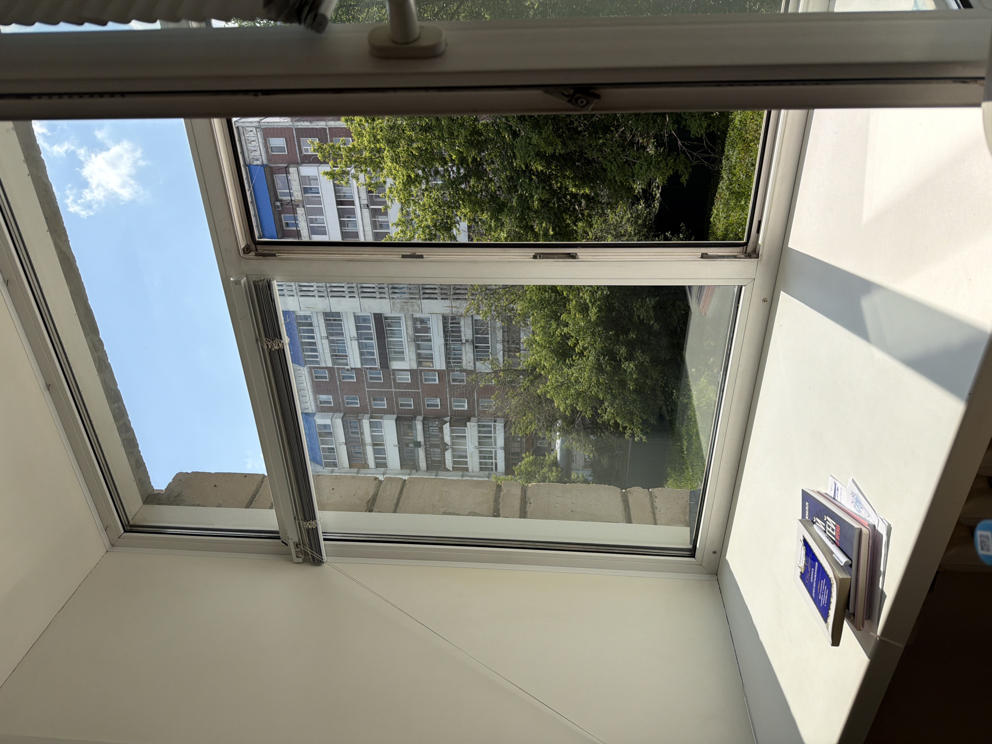 This is an exterior view through an open window, looking out onto a residential area. The primary subjects are two apartment buildings and a dense cluster of green trees. The buildings appear to be older Soviet-era blocks, characterized by their uniform rows of windows and concrete facades. Sunlight streams in, casting strong shadows across the interior sill, suggesting it is daytime. The sky visible in the upper left of the frame is blue with a few white clouds, indicating fair weather.
In the foreground, on the interior windowsill, there is a small stack of books and papers. The top book is partially visible and appears to have a blue cover with some white text. The books are leaning against the window frame.
The perspective suggests the camera is positioned inside an apartment or room, looking outwards. The open window allows for a direct view of the scene outside. The window itself is a modern white frame, and there are blinds partially visible within the frame. The presence of the apartment buildings and trees suggests an urban or suburban environment. The lighting indicates it is likely late morning or early afternoon. No people are visible in the frame. The scene evokes a sense of everyday life in a city.
Some visible text on the book is "ТЕХНИЧЕСКИЙ КОДЕКС" which translates to "TECHNICAL CODE".