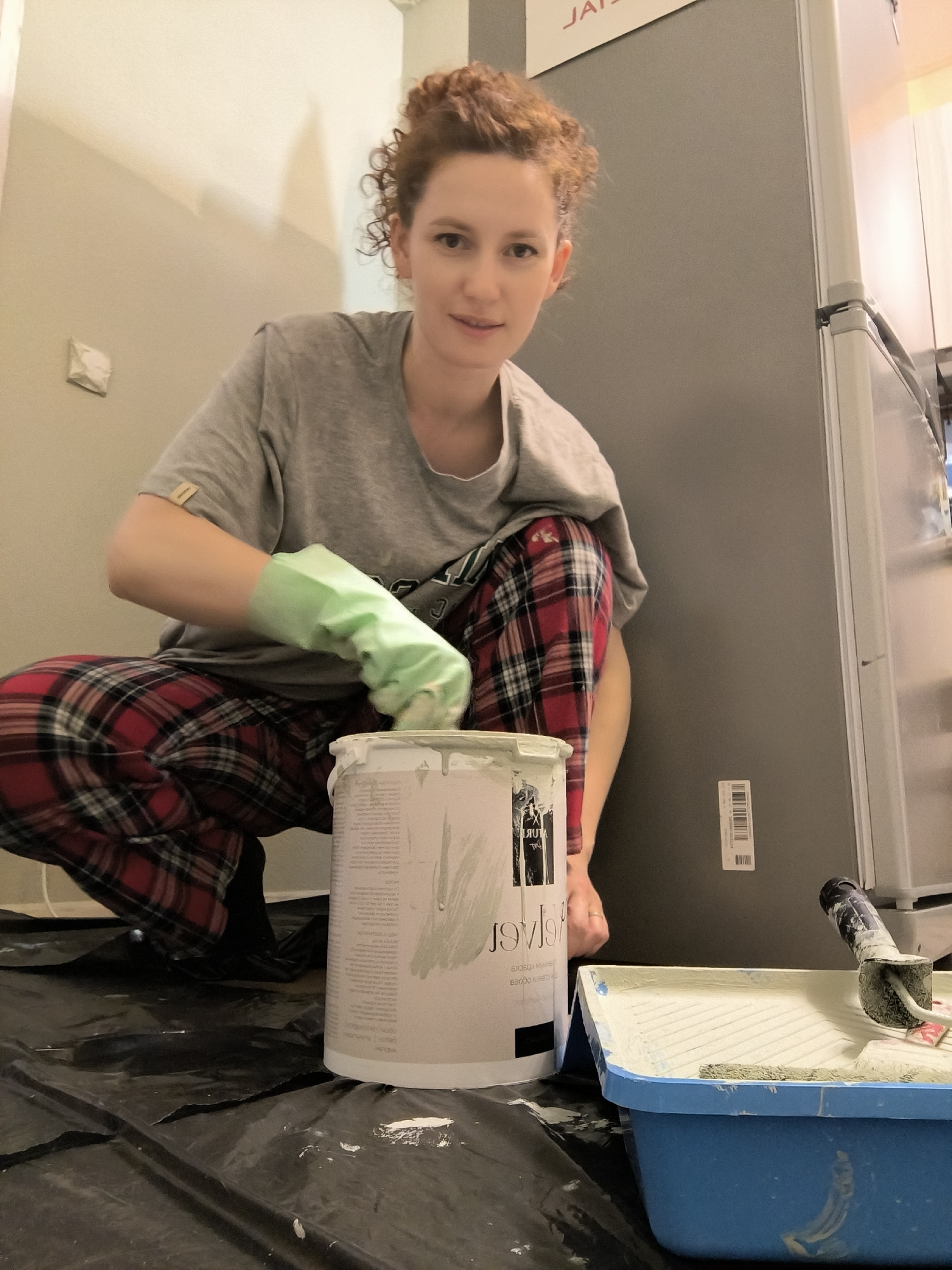 An adult female is captured in a domestic interior setting within Saint Petersburg, Russia, engaged in painting activities. She has reddish-brown curly hair styled upwards, wears a grey short-sleeved t-shirt, red and black plaid pajama pants, and a light green protective glove on her right hand. She is positioned in a crouch or squat, actively stirring light grey paint within a large white cylindrical paint can, which bears Cyrillic text including "ГРУНТ" and "ЭМАЛЬ," consistent with Russian paint products, and is partially stained with paint. Her gaze is directed towards the camera.
To her right, a blue plastic paint tray contains light grey paint and holds a paint roller, which is also coated in paint. The floor beneath the painting area is covered with a black plastic protective sheet, marked with several white paint splatters. The immediate background to the left features a light grey painted wall with a white electrical outlet or switch, partially obscured by white tape. To the right of the individual, a tall, grey, modern refrigerator is visible, affixed with a white barcode label. Above the refrigerator, a partial red sign displaying "JAI" is visible, possibly reversed or part of a larger inscription. The ceiling is light-colored. The scene depicts an ongoing home renovation or maintenance project within a residential space.