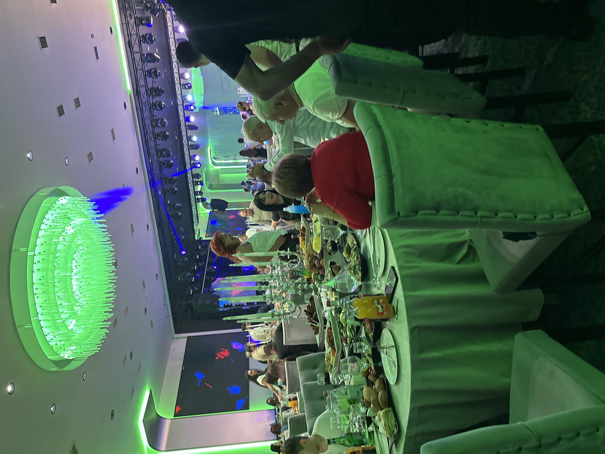 The image depicts a dimly lit banquet hall, likely at the "Aghashen" restaurant in Parakar, Armenia, characterized by vibrant green lighting. A large, modern chandelier with numerous green illuminated rods hangs from the ceiling. The hall is furnished with round tables draped in green tablecloths, surrounded by upholstered chairs, many of which are also covered in green fabric. Several tables are laden with food, including platters of meat, salads, and bread. In the foreground, a table is set with plates, cutlery, glasses, and bottles of orange soda. People are seated at the tables, engaged in conversation and dining. The background features a stage with lighting rigs and a large screen displaying abstract colorful graphics. The overall atmosphere suggests a festive gathering or event.