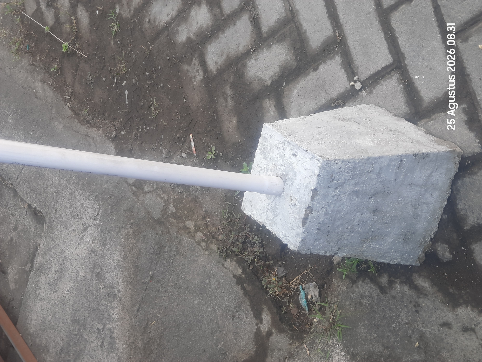 The image shows a white cylindrical pole emerging from a rough, grey concrete base. The base is approximately square and tapered, with a textured surface. The surrounding environment consists of a cracked grey concrete surface on the left and a paved area made of grey interlocking bricks on the right. Small patches of dirt and sparse green vegetation are visible between the concrete and brick surfaces. A metallic brown railing is partially visible in the upper left corner. The date and time "25 Agustus 2026 08.31" are superimposed on the lower right portion of the image. The scene appears to be outdoors, possibly a street or walkway in Sukokerto, Indonesia.