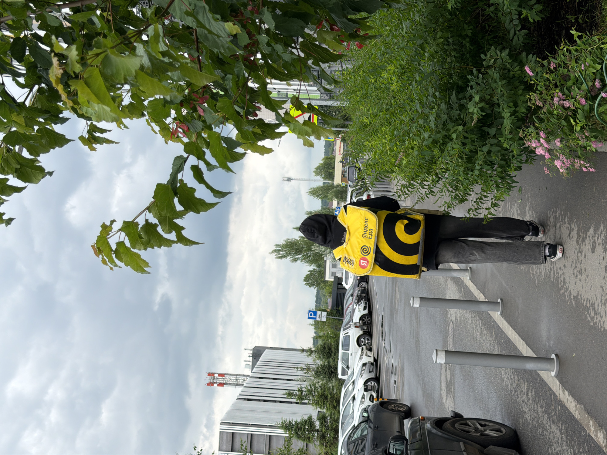 A person walks away from the camera on a wet sidewalk, wearing a black hooded sweatshirt and dark jeans, carrying a bright yellow delivery backpack with a black spiral design and "Yandex.Eda" branding. The person is walking past a line of parked cars in a parking lot. To the left, a modern building with vertical white slats is visible. To the right, lush green bushes with small pink flowers line the sidewalk. The sky is overcast with grey clouds, and there are some trees and utility towers in the background.