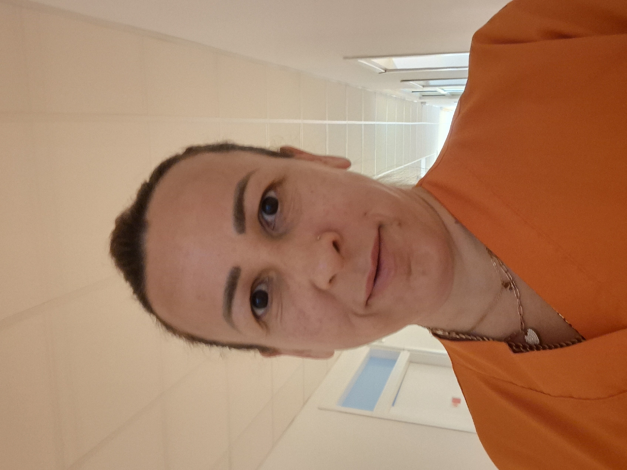 A woman is pictured in a selfie in a hospital hallway. She is wearing an orange scrub top and two necklaces. The hallway is brightly lit with a white tiled ceiling and pale walls. Doors are visible on either side of the hallway.
