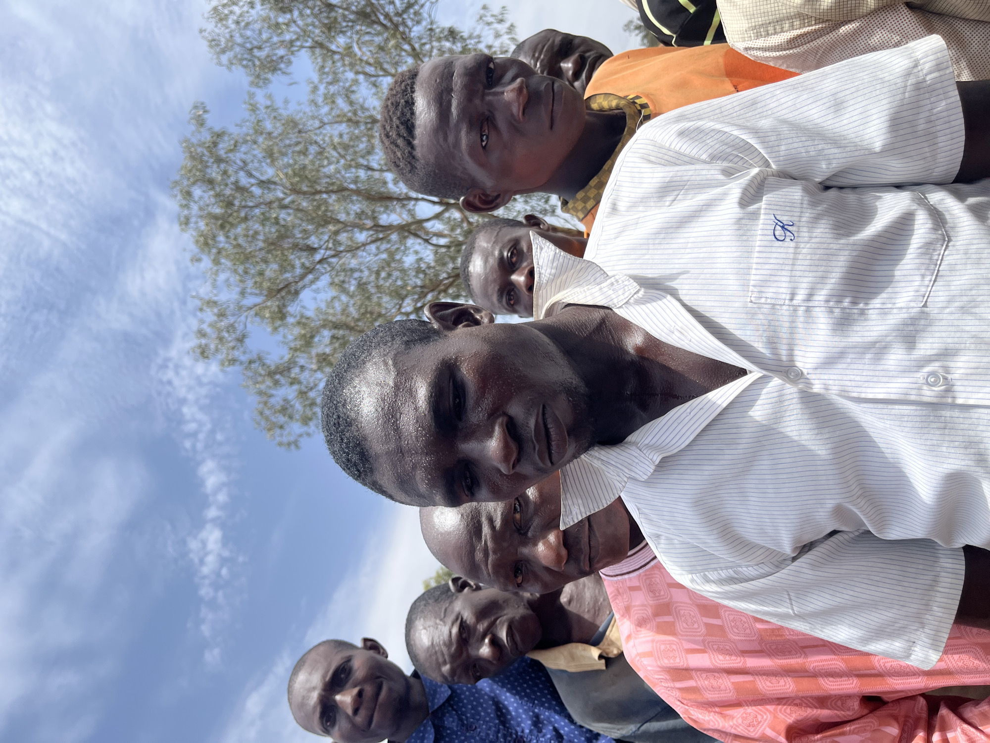 The photograph shows a close-up group of at least six African men in Dutsen Kato, Nigeria. The men are positioned closely together, with their faces prominent in the frame. Their skin appears moist, possibly due to heat or perspiration. Clothing varies, including a white pinstripe shirt with blue embroidery on the pocket, an orange shirt, a pink patterned shirt, and a blue patterned shirt. The background features a blue sky with light clouds and the silhouette of a tree.