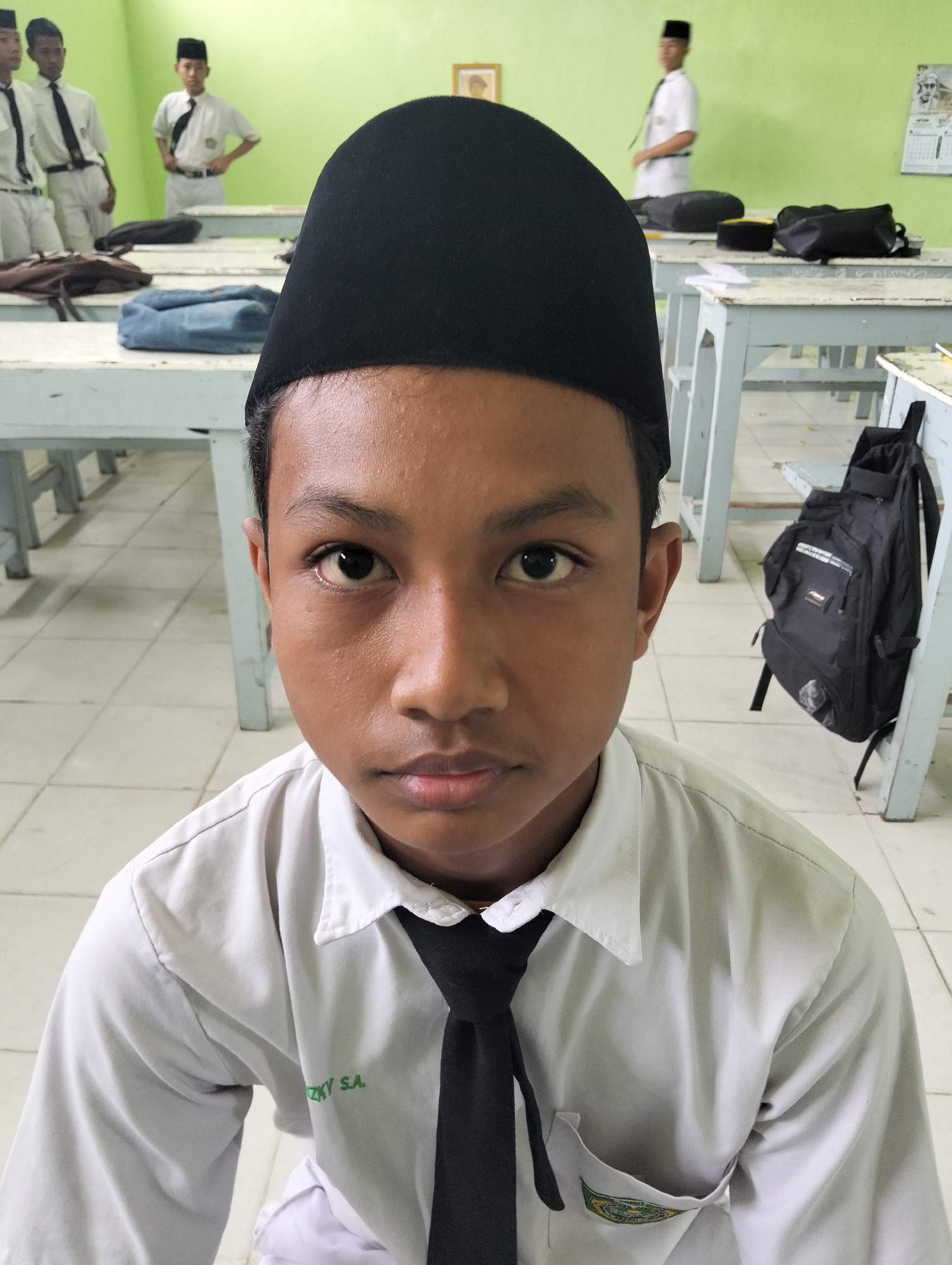A medium shot captures a young Indonesian boy in a classroom setting in Kranji, Indonesia. The boy, centered in the frame, wears a white collared shirt with a black tie and a black peci hat. He looks directly at the camera with a neutral expression. His attire suggests he is a student.
The background reveals a typical classroom environment with rows of light-colored wooden desks and chairs. Other students, also in similar uniforms, are visible in the background, some standing and others out of focus. A black backpack is propped against a desk on the right side of the image. The walls are painted a light green, and a framed picture and a calendar are visible on the wall. The floor is tiled with square, light-colored tiles. The lighting appears to be overhead fluorescent lighting, common in Indonesian classrooms. The overall scene depicts a casual school day.