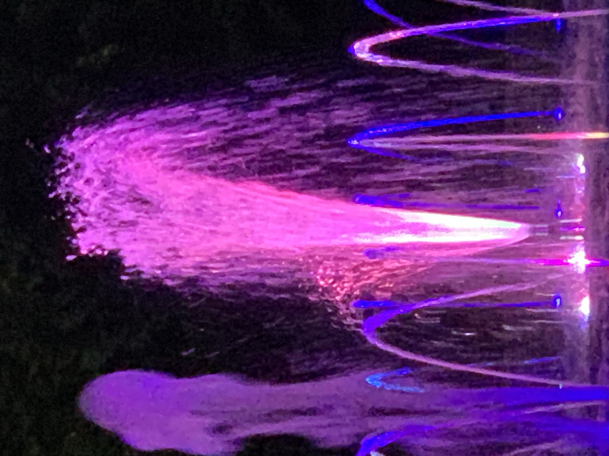 An outdoor night shot captures a fountain illuminated with vibrant pink and purple lights. Several water jets arc upwards, some forming broad sprays and others thin streams. The background is dark and indistinct, suggesting a nighttime setting.