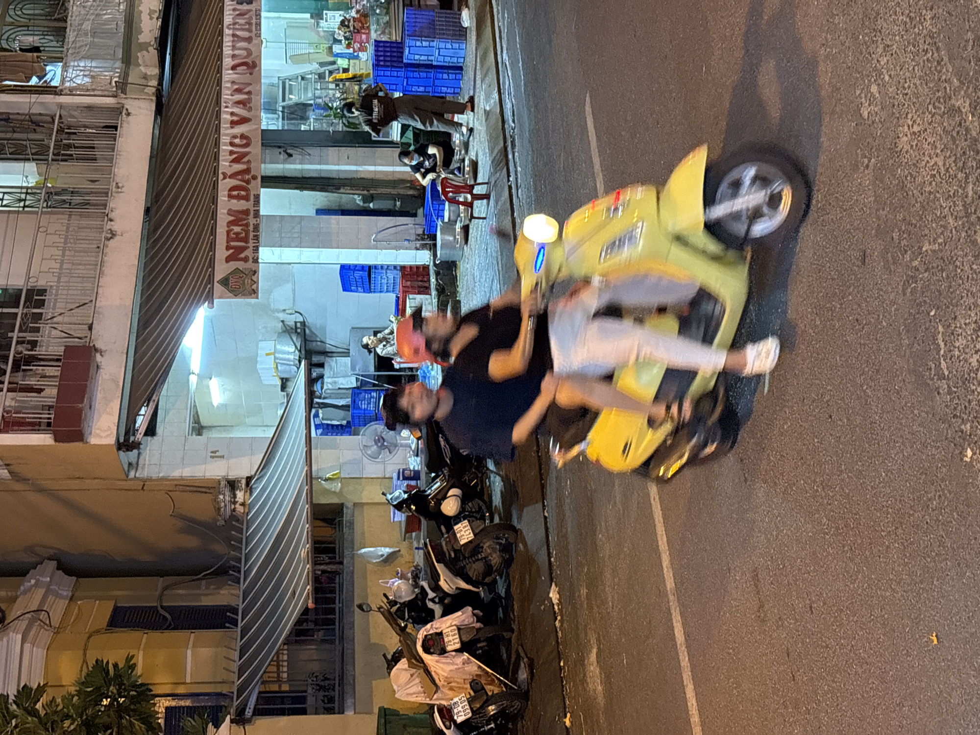 This image depicts a street scene in Nha Trang, Vietnam, at night. The foreground is dominated by a yellow scooter carrying two people. The rider in front, a woman, wears a red cap and white pants, while the passenger behind her, a man, has dark hair and a dark shirt. They are both in motion, with the scooter's headlights illuminated.
In the midground, several parked motorcycles are visible, some covered with white cloths. The background features the exterior of a building with a striped awning and a sign that reads "NEM ĐẶNG VĂN QUYÊN". There are storefronts with blue crates stacked outside. Several individuals are visible in the background, some standing and others appearing to be working or interacting. The overall environment suggests a commercial area of the city.