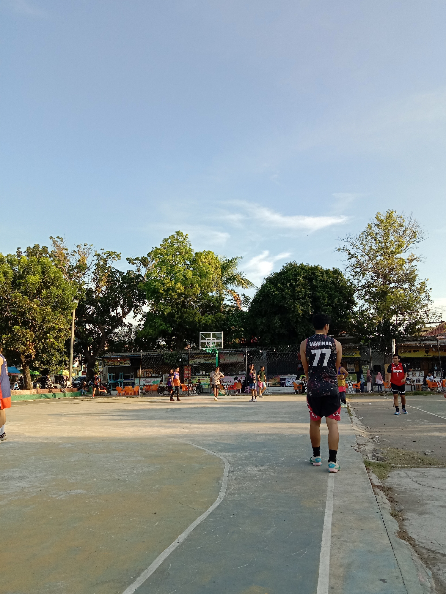 The photograph depicts a public basketball court in Lhokseumawe, Indonesia. Several individuals are playing basketball, with one player wearing a jersey with the number "77" and the name "MARINDA" on the back. Another player wears a red jersey with the number "1" and "INDONESIA" printed on it. In the background, there is a structure with orange chairs, and people are sitting and standing around it. Trees surround the court, and a blue sky with some clouds is visible above. The court surface is grey and marked with white lines.