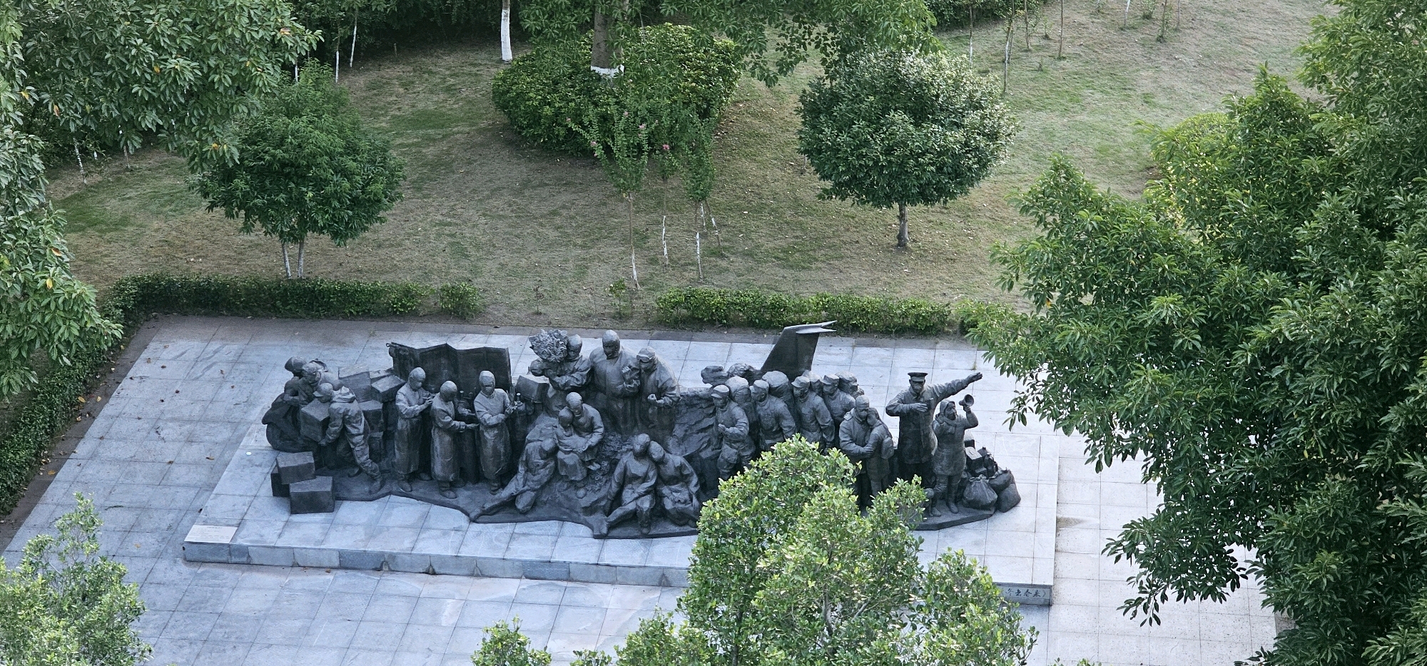 The image shows a bronze sculpture depicting a group of people in historical attire, seemingly engaged in a collective activity or commemoration. The sculpture is situated outdoors on a paved plaza, surrounded by manicured greenery and trees. The scene is viewed from a high vantage point, looking down onto the sculpture. The overall impression is of a memorial or monument, possibly commemorating a significant event or group of individuals. The location is identified as the Chongqing High-tech Zone, China, suggesting a public space within an urban or suburban setting.