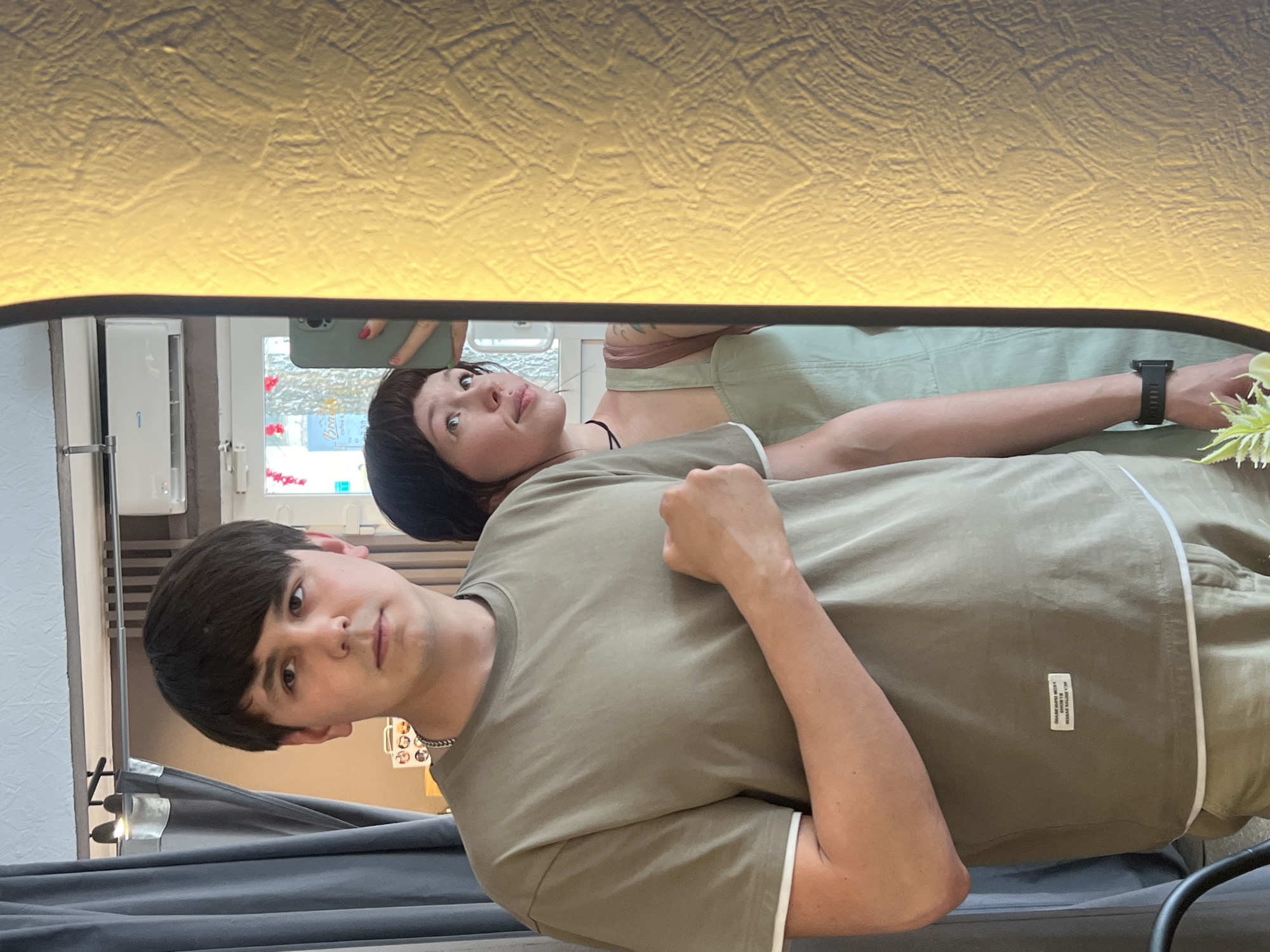 A reflected image in a mirror shows two people. The mirror is framed with an opaque gold material at the top and black material on the sides. The top frame is illuminated with a warm light source.
A man is positioned in the lower portion of the mirror's reflection. He is looking directly at the viewer and his arm is raised. He has dark hair, fair skin, and is wearing a light brown T-shirt with white trim at the edge of the sleeve. A small white tag is visible on the lower right side of his shirt. A silver chain necklace is visible.
Behind him, a woman is visible in the upper portion of the mirror's reflection, looking upwards. She has dark hair and is wearing a light green jumpsuit. Her left arm is extended behind the man, with a smartwatch visible on her wrist.
The background includes reflections of windows, wall surfaces, and a radiator. A green phone is being held by the woman. A light fixture with three bulbs is visible.
The location is possibly Chaikovsky, Russia, based on information provided.