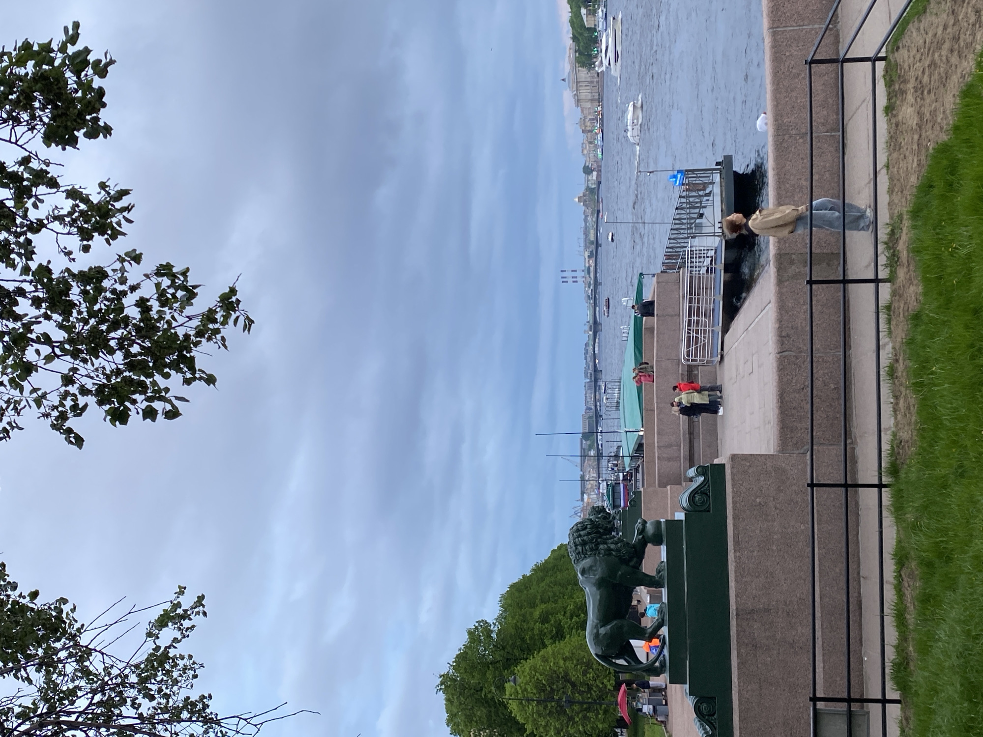 The image captures a section of a river embankment in Saint Petersburg, Russia. A bronze lion sculpture stands prominently on a pedestal to the left of the frame, with green trees behind it. The Neva River stretches into the distance, populated by several boats and framed by buildings along the distant shoreline. Several people are visible along the embankment, including a person in a beige jacket standing near a protective railing and other individuals further down the walkway. A section of black metal fencing runs along the edge of the embankment, alongside a patch of grass. The sky is overcast with a layer of grey clouds.