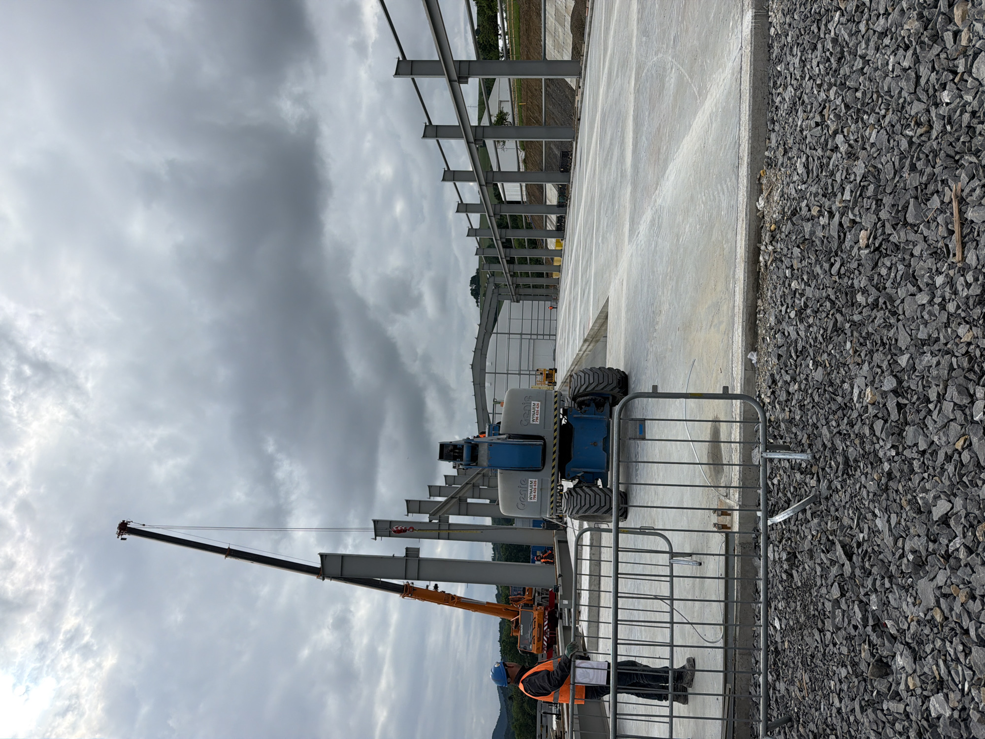 The image depicts a construction site with a large, unfinished building. In the foreground, a person wearing a hard hat and a high-visibility vest stands near a metal barrier. Behind them, a blue scissor lift is parked on a concrete surface. Further in the background, a large orange crane is lifting a steel beam into place for the building's structure. The sky is overcast with heavy clouds. The scene is consistent with ongoing construction work at the stated location in Stróża, Poland.