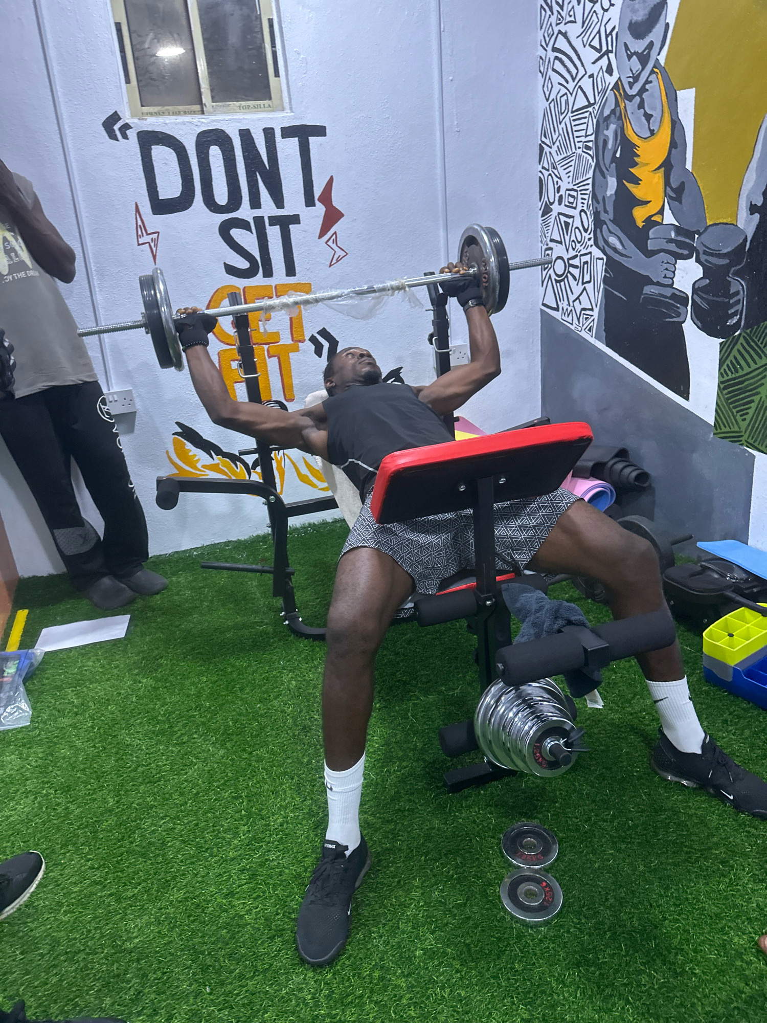 The image depicts an indoor gym with artificial turf flooring in Badagry, Nigeria. A man is performing a bench press exercise with a barbell. He is lying on a black and red weight bench, wearing a black tank top, patterned shorts, white socks, and black athletic shoes. His hands are gloved as he grips the barbell, which has silver weight plates on either side. To his left, another person, partially visible, is standing and wearing a grey t-shirt and black tracksuit pants. The wall behind them is decorated with graffiti, including the words "DON'T SIT" and "GET FIT," as well as a mural of a muscular figure holding dumbbells. Various gym equipment, including dumbbells and weight plates, are scattered around the room. A yellow and blue plastic storage bin is visible in the background.