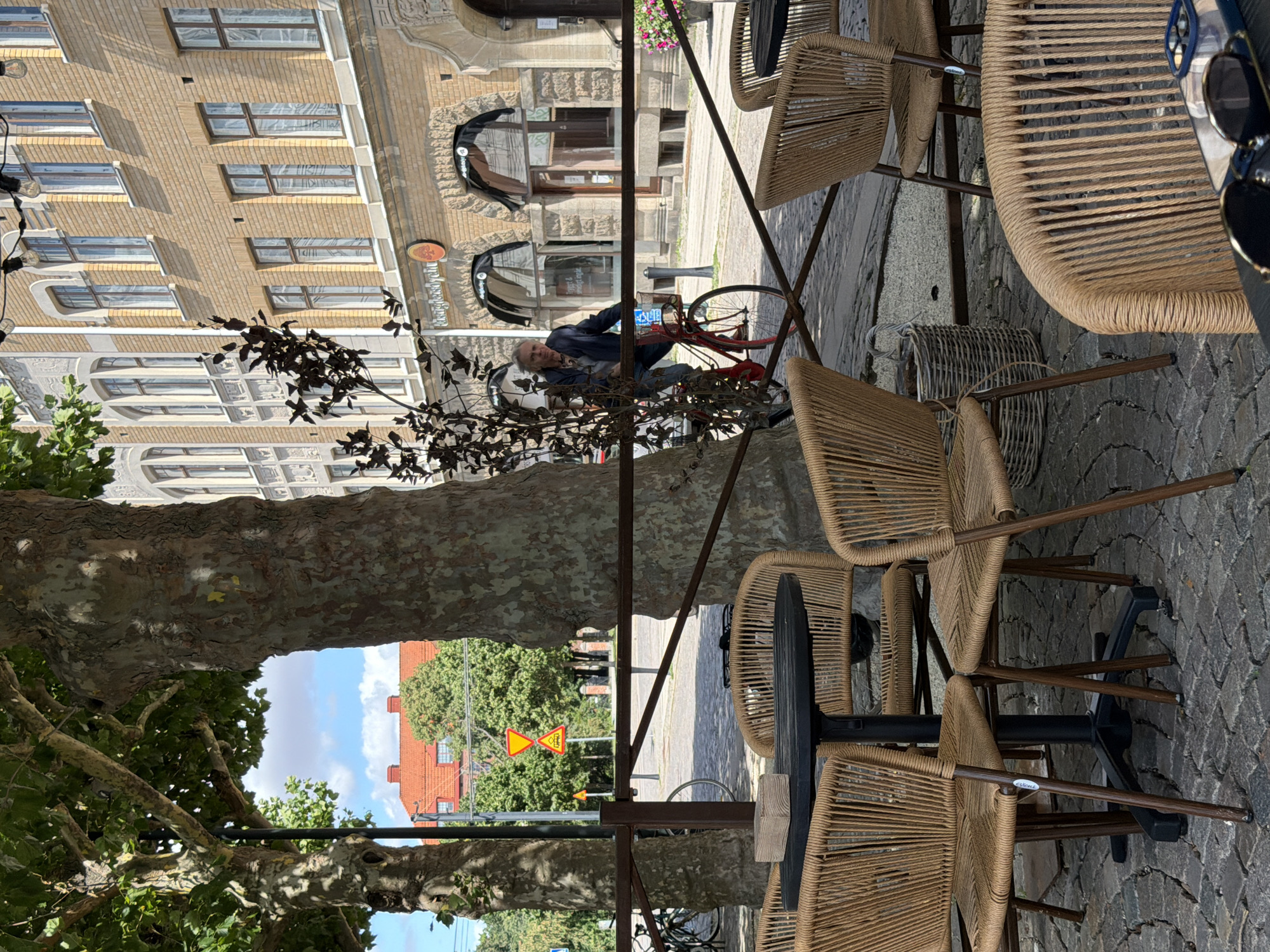 This is a medium shot photo taken outdoors at a cafe with a view of the street in Lund, Sweden. In the foreground, there are several outdoor tables and chairs made of woven rope material. A black round table in the center has a small wooden object on it. Behind the tables and chairs, a brown metal railing separates the cafe seating area from the sidewalk.
Beyond the railing, a man with gray hair is seen riding a red bicycle. He is wearing a dark blue jacket and jeans. The bicycle has a basket on the handlebars and a red frame. The man is looking towards the left of the frame.
In the background, there is a brick building with multiple windows and architectural details. A street sign with yellow warning symbols is visible on the left side of the street. Trees with green leaves are present, casting shadows on the ground. The overall atmosphere suggests a daytime scene on a city street.