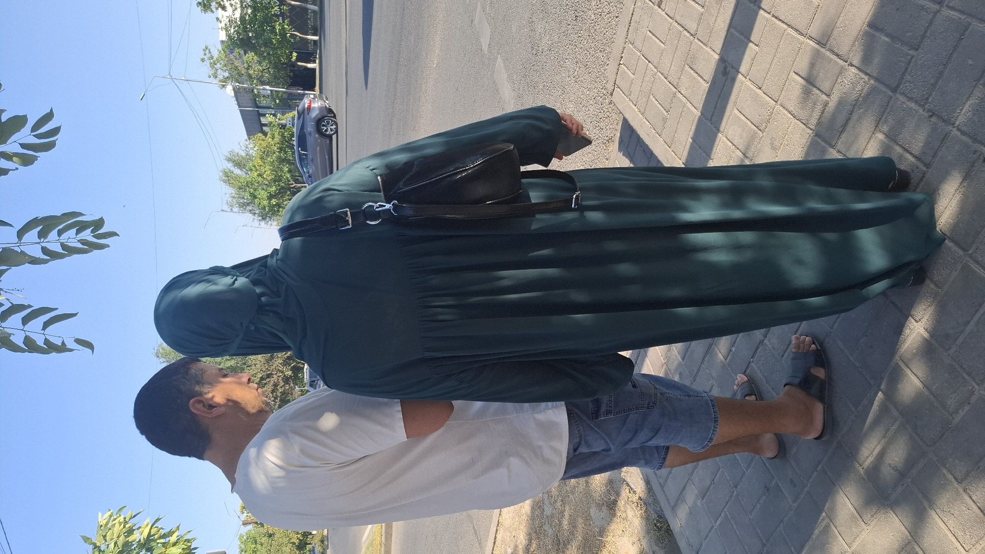 This image depicts two individuals standing on a paved sidewalk adjacent to a road in the city of Shymkent, Kazakhstan. The person on the left, a man wearing a white t-shirt and denim shorts, is looking down, possibly at a phone. The person on the right is wearing a dark green abaya and hijab and is holding a phone in their right hand. A black handbag is slung over their shoulder. The road features a passing car, streetlights, and trees lining the sidewalk. The scene appears to be outdoors during daylight hours.