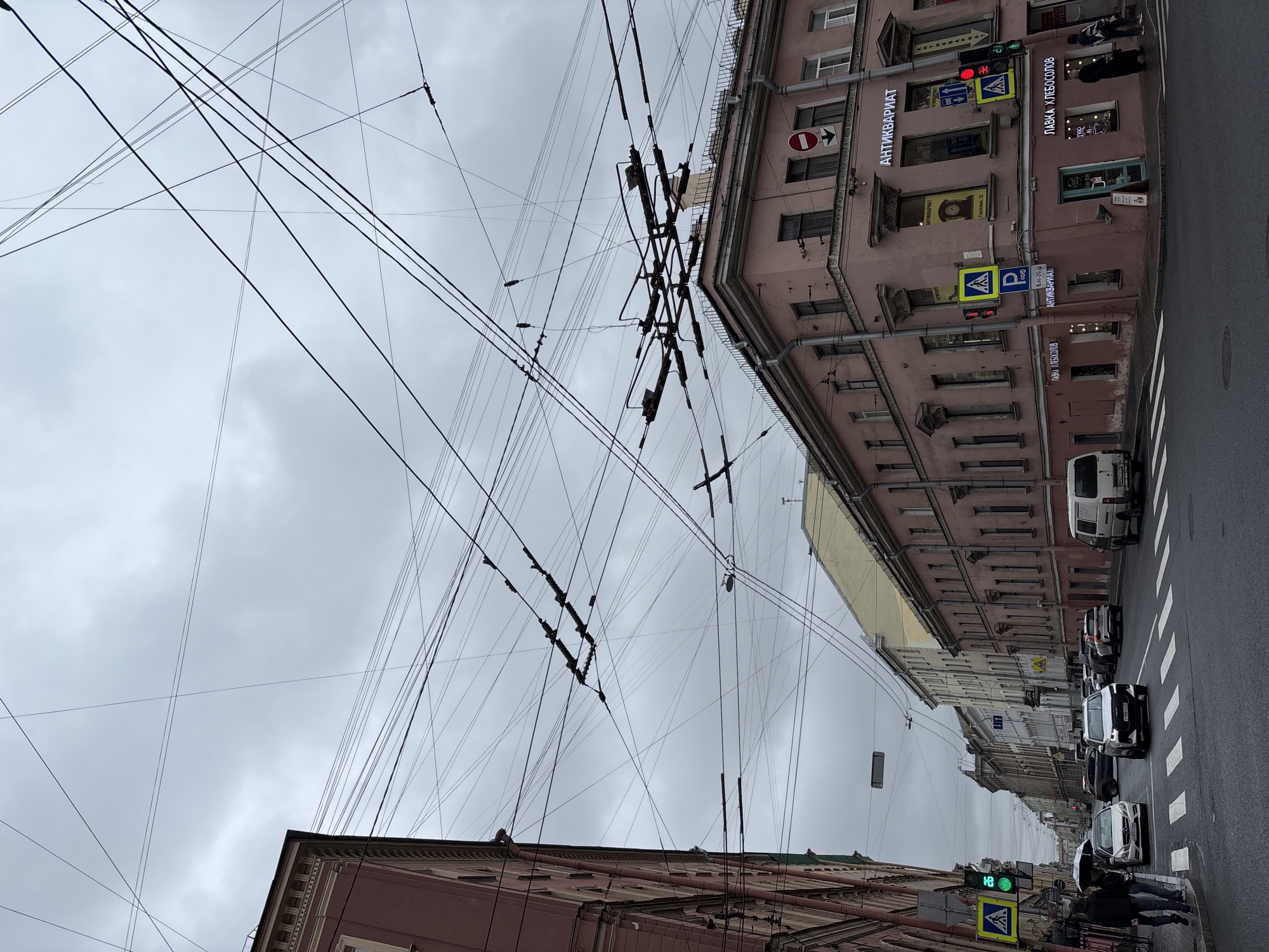 The image shows a street scene in Saint Petersburg, Russia, characterized by overhead power lines crisscrossing the sky. Buildings line the street, featuring architectural details such as windows, gutters, and signs in Russian. The building on the right has signs for "Antikvariat" and "Lavka Hlebosolov". Pedestrian crossing signs are visible. A traffic light shows both red and green signals. Several vehicles are on the road, including cars and a van. Pedestrians can be seen on the sidewalk. The sky is overcast.