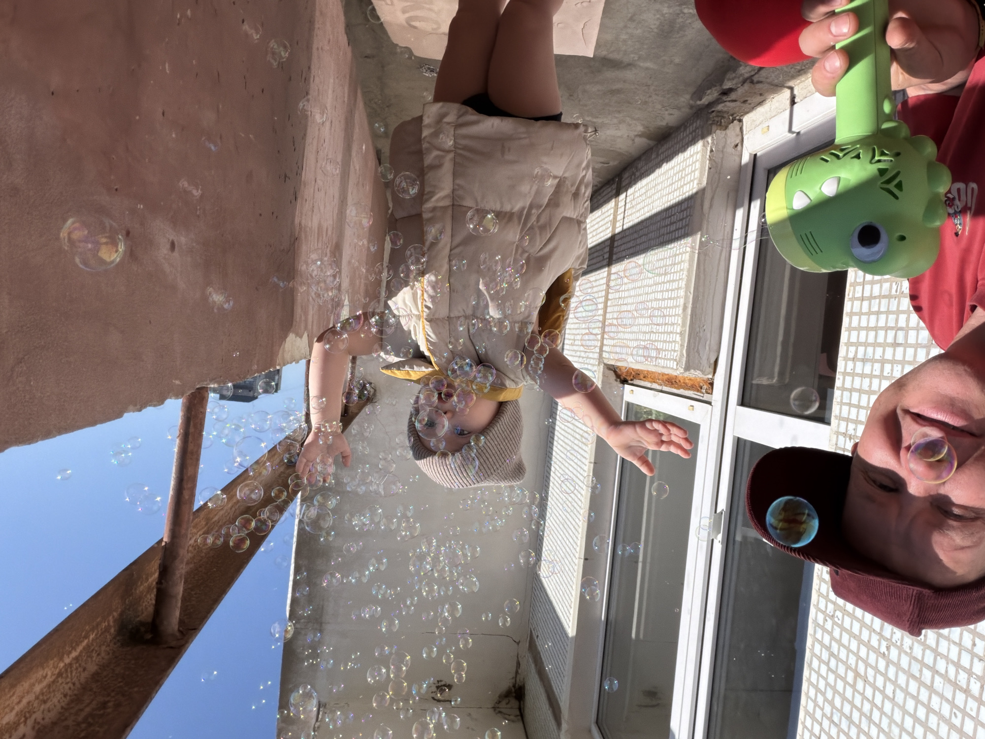 A rotated low-angle shot shows a child playing with bubbles with an adult, likely in Chaikovsky, Russia. The child, wearing a vest and a hat, is reaching out to catch the bubbles. Bubbles are floating in the air around the child and the adult. The adult is holding a bubble-making toy. The background includes a building exterior with white tiles and a window. The sky is visible.