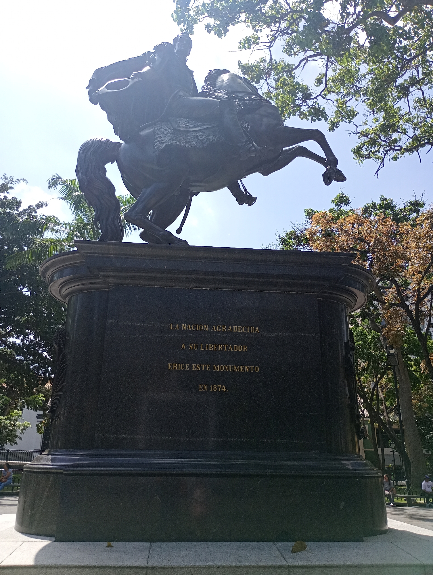 The image depicts a bronze equestrian statue mounted on a dark stone pedestal. The inscription on the pedestal reads "LA NACION AGRADECIDA A SU LIBERTADOR ERICE ESTE MONUMENTO EN 1874". Trees surround the statue, with glimpses of a clear sky. The photo appears to be taken from a low angle. The location is likely Plaza Bolívar in Caracas, Venezuela, where the equestrian statue of Simón Bolívar, erected in 1874, is located. There are people sitting on benches in the park surrounding the statue.