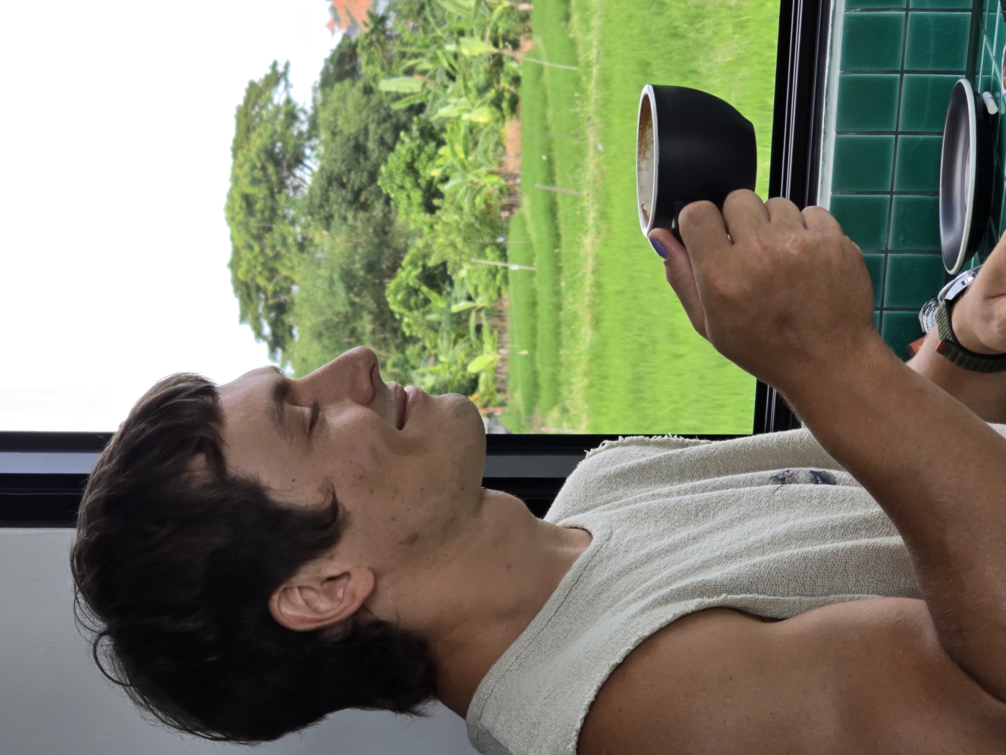 A man with brown hair is shown in profile, holding a black cup and saucer in his right hand, in an indoor setting. The man is wearing a light-colored sleeveless shirt. He is looking at the cup. Outside the window behind him is a view of green rice paddies and trees. The location is North Kuta, Indonesia. Green tiles are visible on the wall to the right of the window, and a watch on the man's left wrist is also visible.