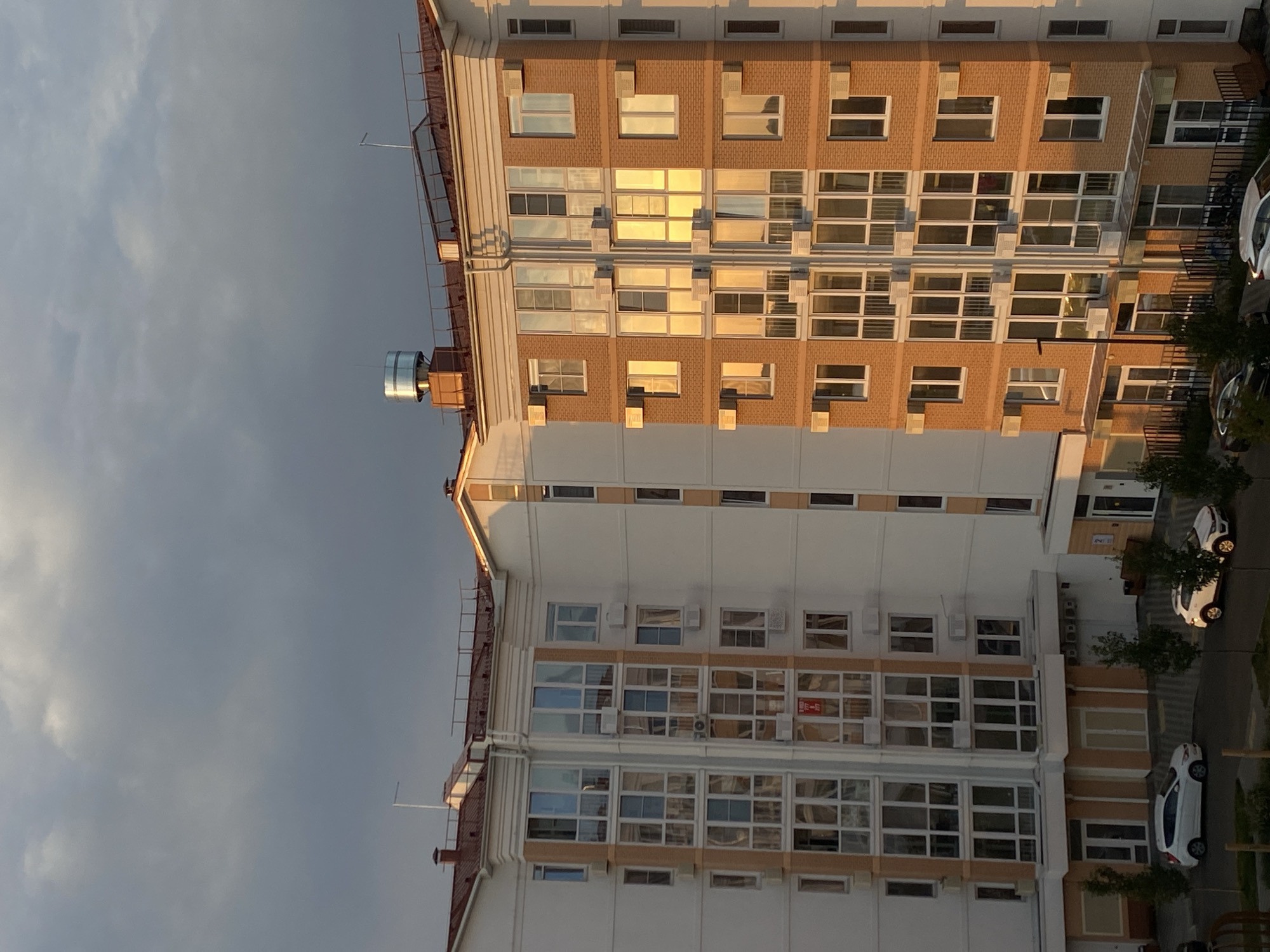 The image shows a residential apartment building in the city of Dinskaya, Russia. The building is modern with white and orange brick facades, numerous windows, and balconies. Several air conditioning units are visible mounted outside the windows. The sky is overcast with dark clouds. In the foreground, there are cars parked on the street. The scene appears to be captured during the late afternoon or early evening, as indicated by the warm lighting reflecting off the windows.