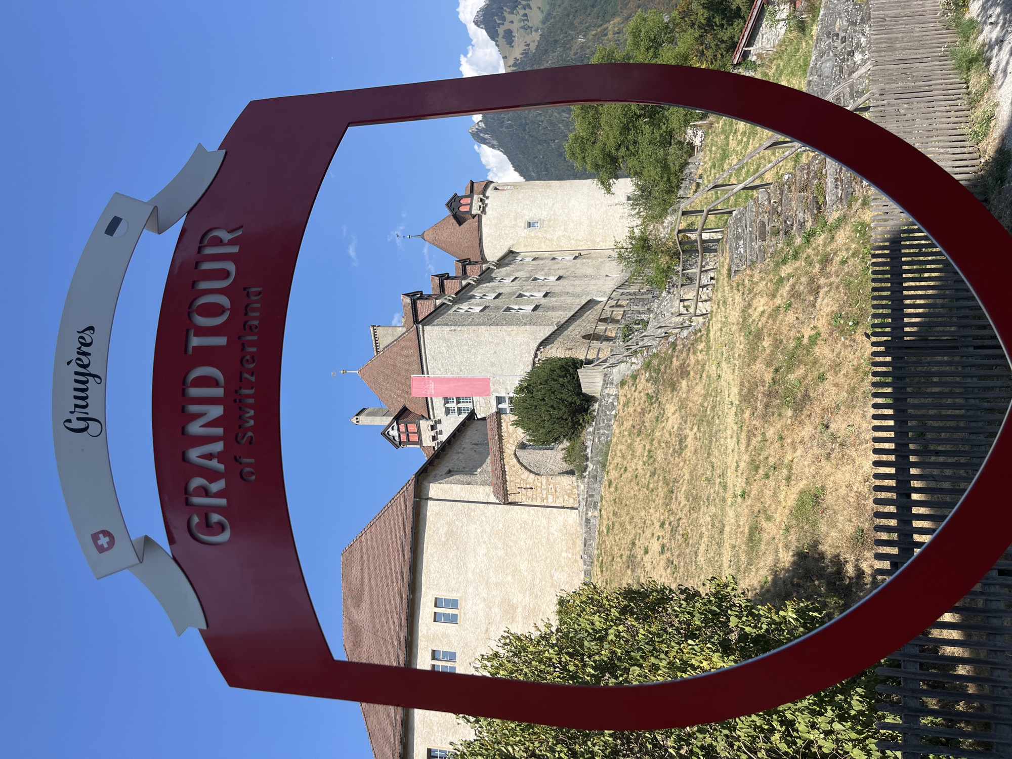 The image is an outdoor shot of the Gruyères Castle in Gruyères, Switzerland. The castle is visible through a large, red, shield-shaped cutout sign that reads "GRAND TOUR of Switzerland" and "Gruyères". The castle is a stone building with a red-tiled roof and several towers, set on a grassy hill. A flag is visible on one of the towers. The sky is clear and blue with some clouds. In the foreground, there is a wooden fence. The scene appears to be a tourist attraction on a sunny day.