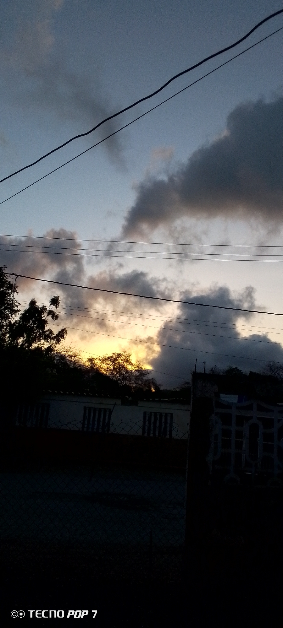 The image depicts a dusky sky over a residential area. Several power lines crisscross the frame horizontally. Dark, billowing clouds are present in the sky, with a sliver of yellow light peeking through them, suggesting the setting sun. Below the sky, silhouetted trees and foliage are visible, with a low-rise building featuring barred windows behind a chain-link fence. A decorative metal gate and a concrete post stand to the right of the frame. The bottom left corner of the image displays a watermark: "TECNO POP 7". The overall impression is of a twilight scene in a somewhat overcast urban or suburban environment.