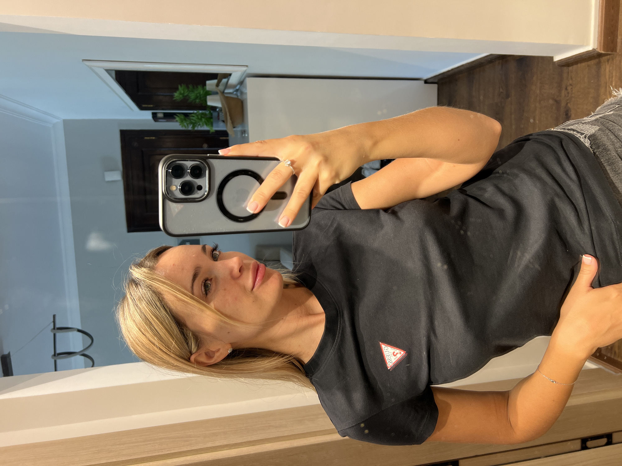 In Sharm El Sheikh, Egypt, a young Caucasian woman with blonde hair is taking a mirror selfie. She is wearing a black t-shirt with a red and white Guess logo on the left chest, and grey denim shorts. She has a ring on her left ring finger and a delicate bracelet on her left wrist. Her phone, which she is using to take the picture, has a clear case with a black MagSafe ring. The background suggests an indoor setting, possibly a hallway or dressing area, with a large mirror reflecting a door and some potted plants. The lighting is moderate, highlighting the woman and her immediate surroundings.