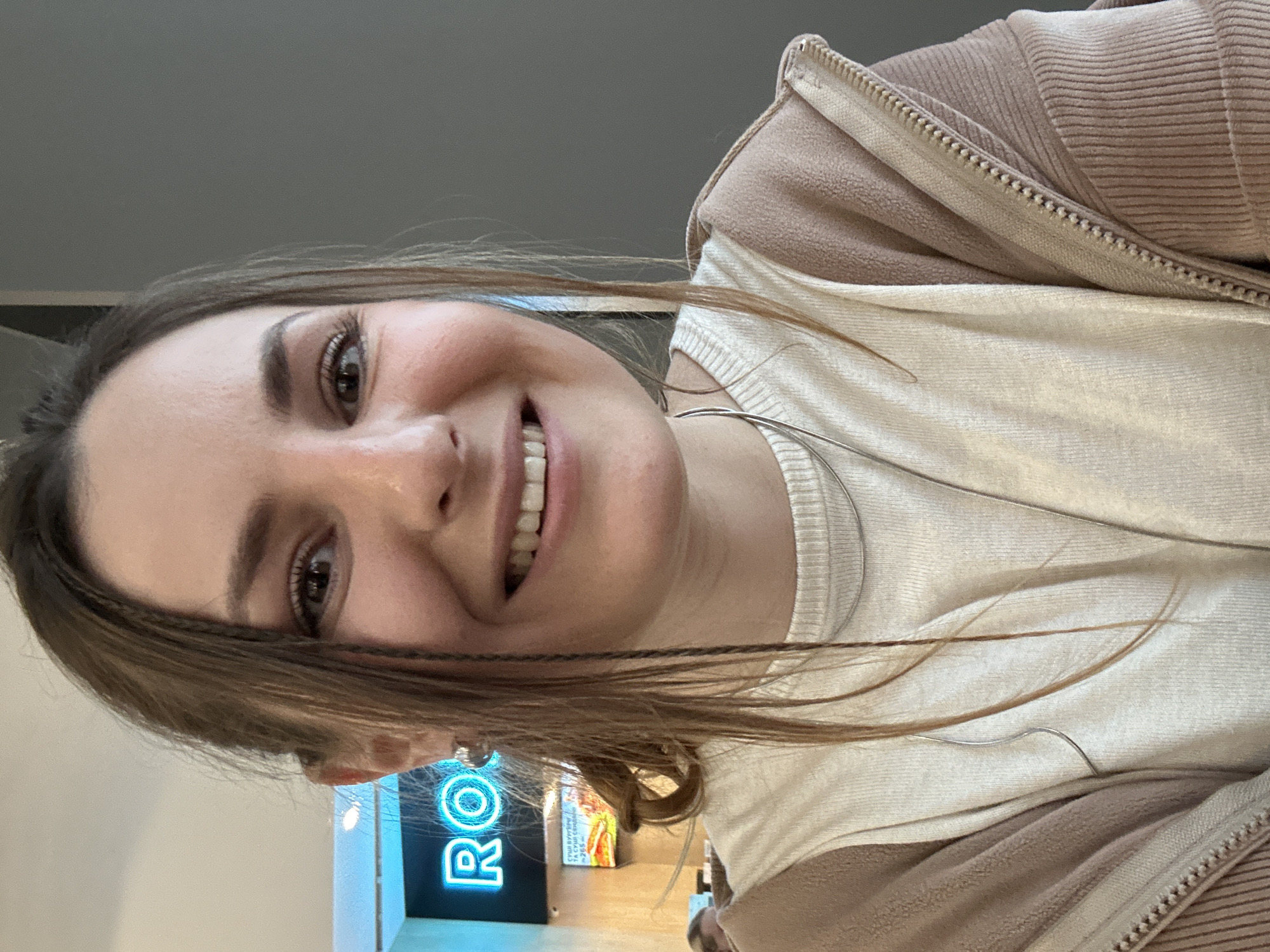 The image is a selfie of a young woman with brown hair smiling at the camera. She is wearing a light-colored sweater under a brown zip-up jacket and a silver necklace. The background appears to be indoors, possibly a store or public space, with a blue neon sign that reads "RO". The overall lighting is bright and appears to be a part of Unknown. The specific location in the city of Unknown, Unknown cannot be determined from the image.