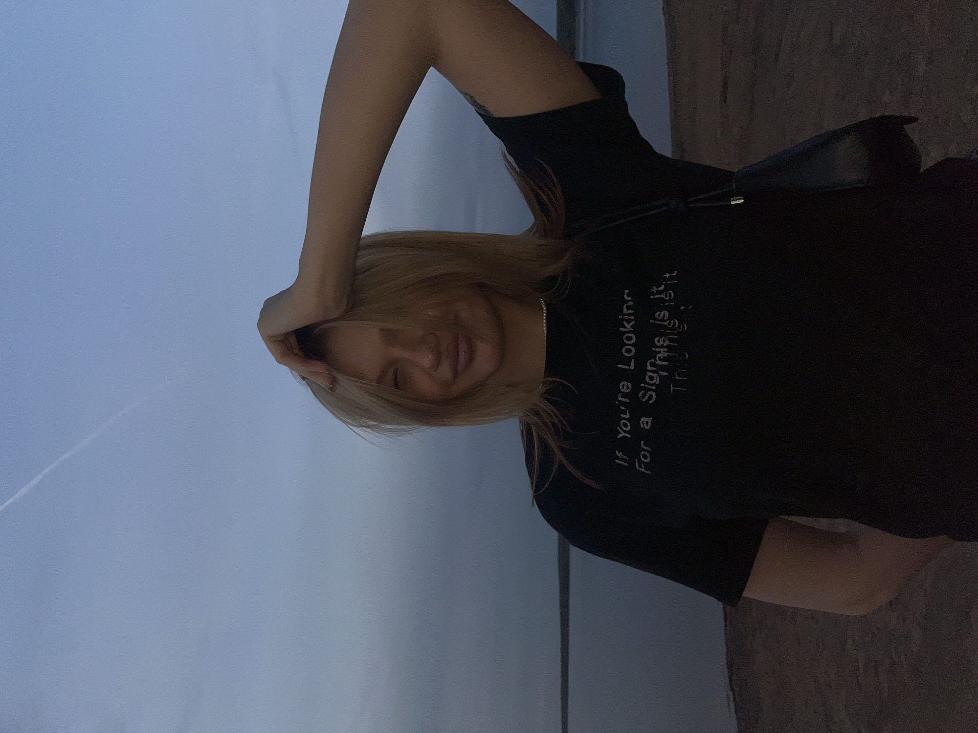 A person with light blonde hair is captured in a medium shot, set against a muted outdoor backdrop of sky and what appears to be a body of water. The person is wearing a dark-colored T-shirt with the printed text "If You're Looking For a Sign, This is It." The individual's right arm is raised above their head, with the hand resting near their forehead. The person is looking into the camera with a slight smile. A small dark purse is visible on their shoulder. The shot has a tilted perspective, which is disorienting to the scene. Based on the provided information, the location is Chaikovsky, Russia.