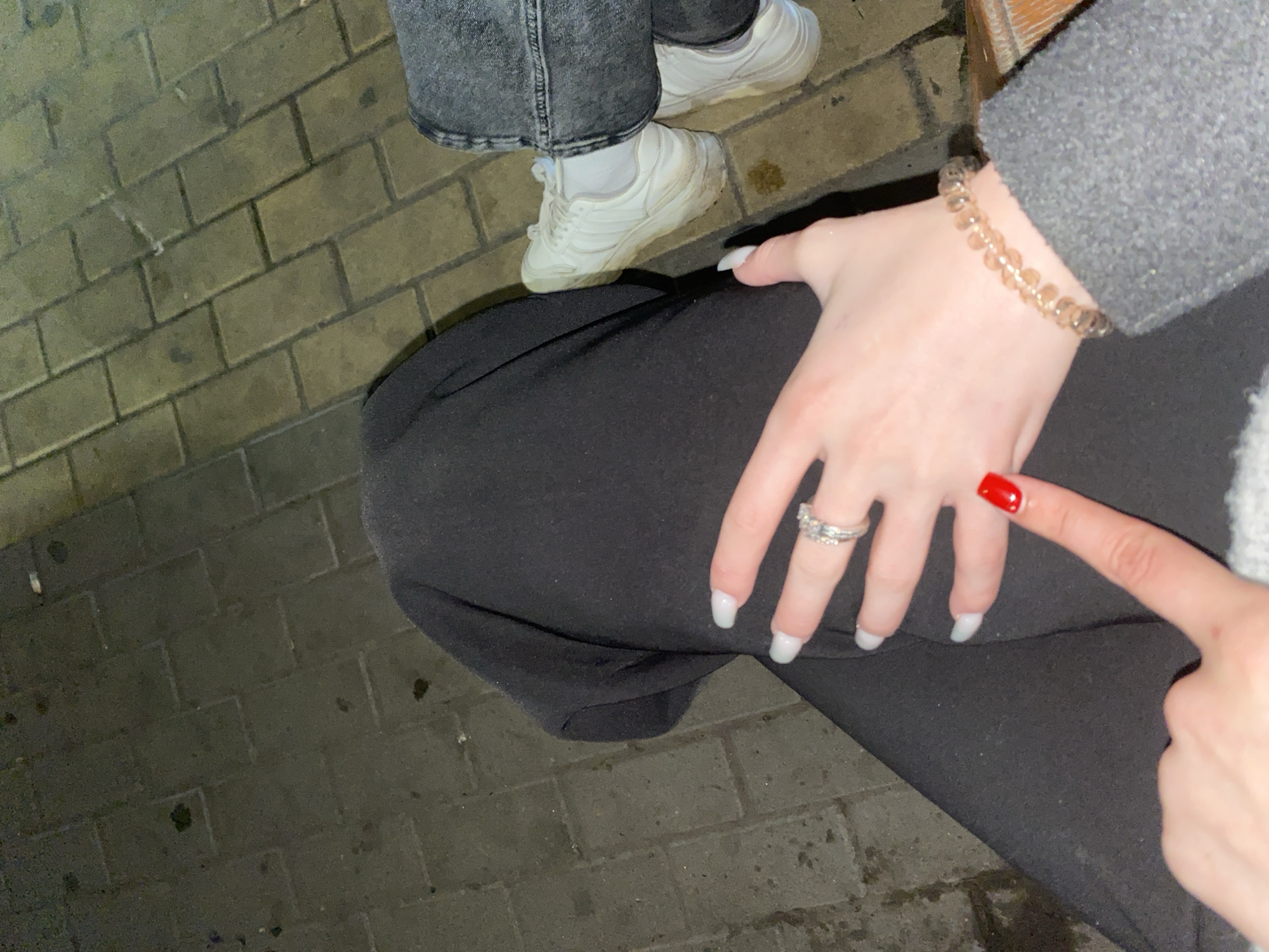 The image captures a close-up, downward perspective of two individuals in an outdoor setting, likely during the evening or night due to the dim lighting. The ground is paved with rectangular, dark-toned brick pavers, which appear slightly damp in certain areas.
One person is partially visible in the upper left, wearing grey denim jeans and white sneakers, standing on the brick pavement. The other individual is largely out of frame, seen only from the waist down and their hands. This person is wearing black trousers and a grey top or jacket.
The primary focus is on the hands of the person wearing black. One hand, adorned with long, white-polished fingernails and a silver-colored ring on the ring finger, rests on their black pants. A light brown, multi-faceted beaded bracelet is on this wrist. Another hand, with a bright red-polished index finger, is pointing directly at the ring, suggesting an act of showcasing the jewelry, perhaps an engagement or a new purchase.
No specific location identifiers are visible apart from the generic brick pavement. There is no visible text within the image. The overall impression is a personal moment centered around the ring.