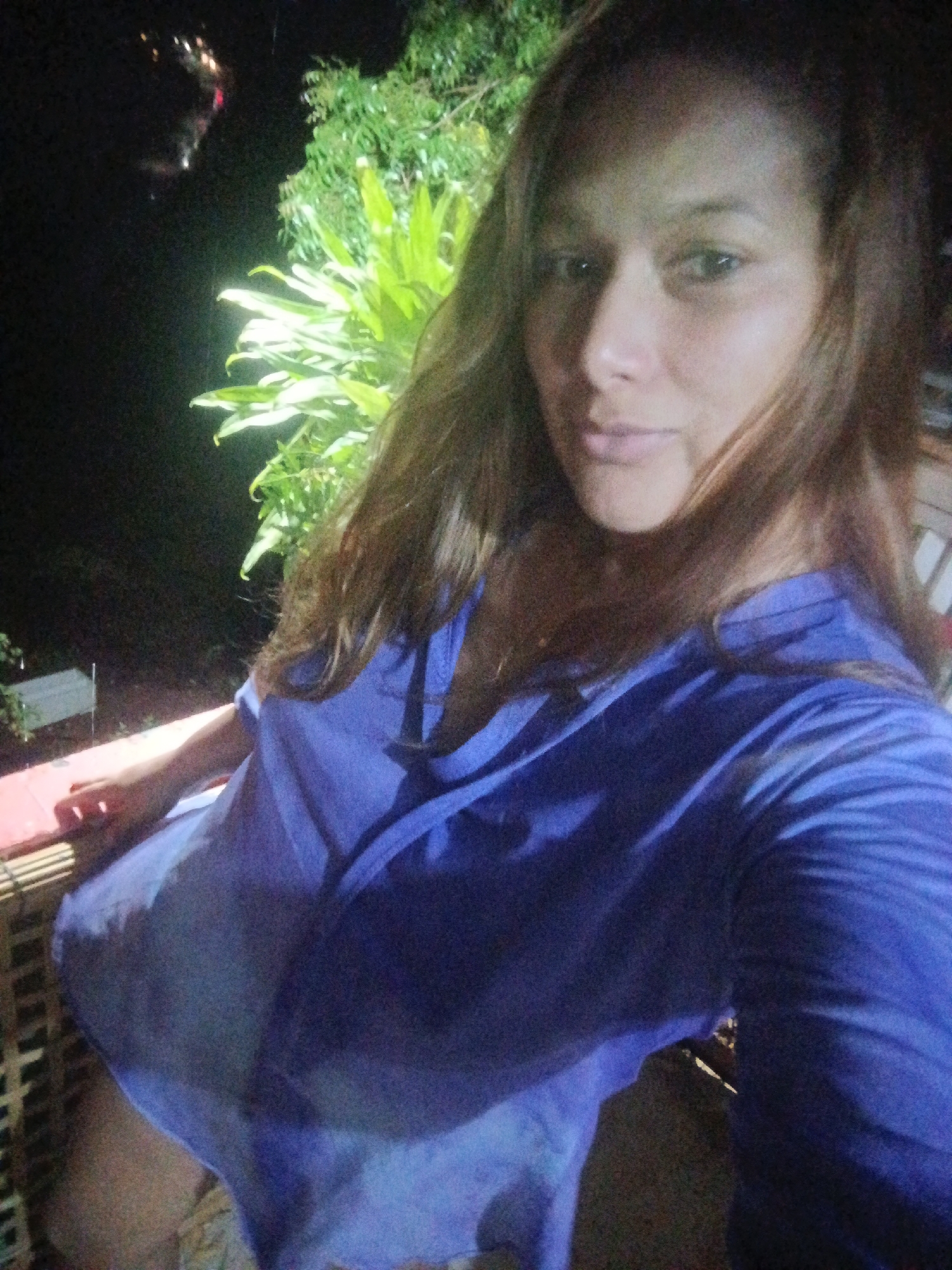 A medium-shot photograph shows a woman with long brown hair, wearing a blue shirt with a deep V-neck, standing on a balcony. Behind her are green plants illuminated by a light source. In the background, it's dark, suggesting it's nighttime. The location is Unknown, Unknown. The woman is facing the camera with a slight smile. The balcony appears to be constructed of bamboo.