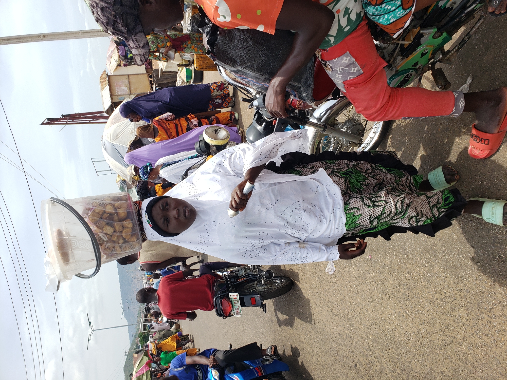 A woman in a white dress and headscarf, carrying a container of fried goods on her head, stands in a street in Lokoja, Nigeria. Other people are visible, including one on a motorcycle. The street has motorcycles and tents. The scenery behind the street includes mountains and clear sky.