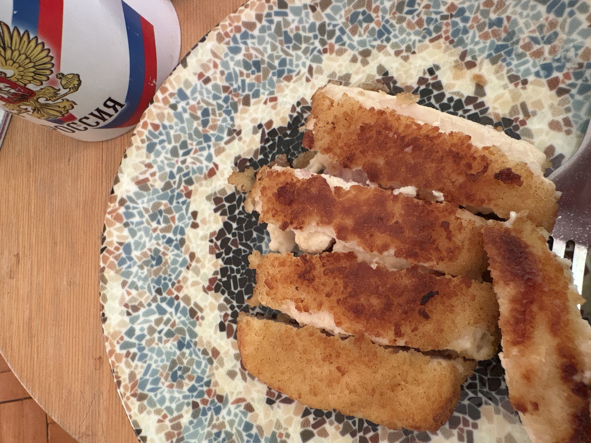 A close-up shot shows a plate of sliced, breaded and fried food next to a mug on a wooden table. The plate is mosaic-patterned with blue, brown, and beige tiles. On the plate, a piece of fried food appears to have been sliced into four pieces, revealing a white interior. To the right of the slices, another piece is visible, held by what looks like the tines of a fork. To the left of the plate, a portion of a white mug is visible with the Russian flag colors and the word "Россия" written on it. The setting appears to be indoors on a wooden surface. No people are visible in the image. The location in the city of Novoe Devyatkino, Russia is inferred from the mug featuring Russian text and emblems.
