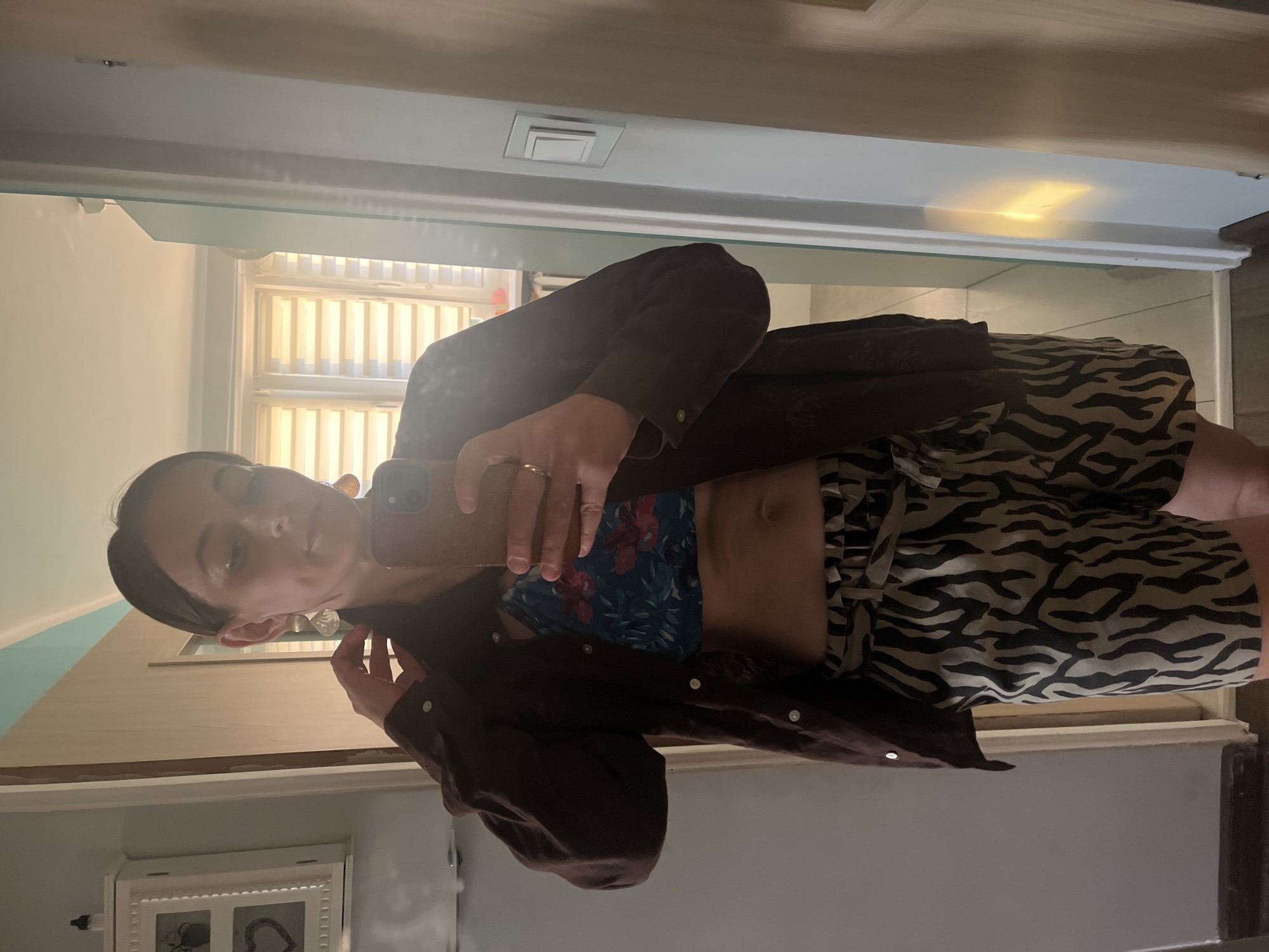 A woman is standing in front of a mirror and taking a selfie. She is wearing a brown unbuttoned shirt over a floral print crop top and zebra print shorts. The woman has a ring on her right hand and is wearing large earrings. The background shows a room with wooden floors and walls, a window with blinds, and a cabinet.