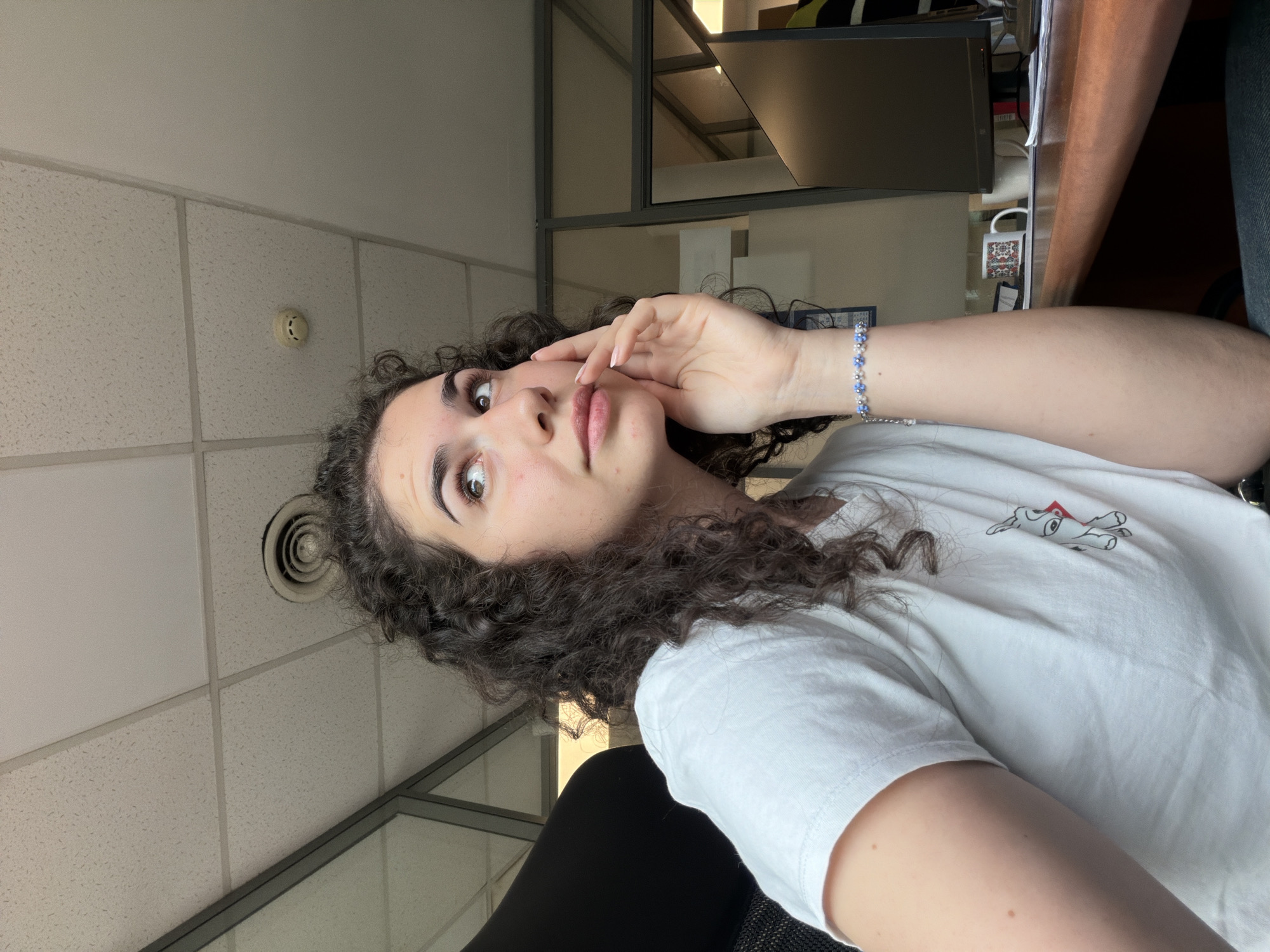 A young woman with curly dark hair is seated at a desk in an office environment. She is wearing a white t-shirt with a small graphic on it and a beaded bracelet on her left wrist. Her right hand is resting on her cheek, and she is looking upwards and to the right. The office has a dropped ceiling with fluorescent lighting and what appears to be a smoke detector. Behind her, glass partitions separate office spaces, and a computer monitor is visible on the desk. The overall impression is that of someone deep in thought or contemplation while at work, possibly in an office located in Sofia, Bulgaria, given the context of the prompt.