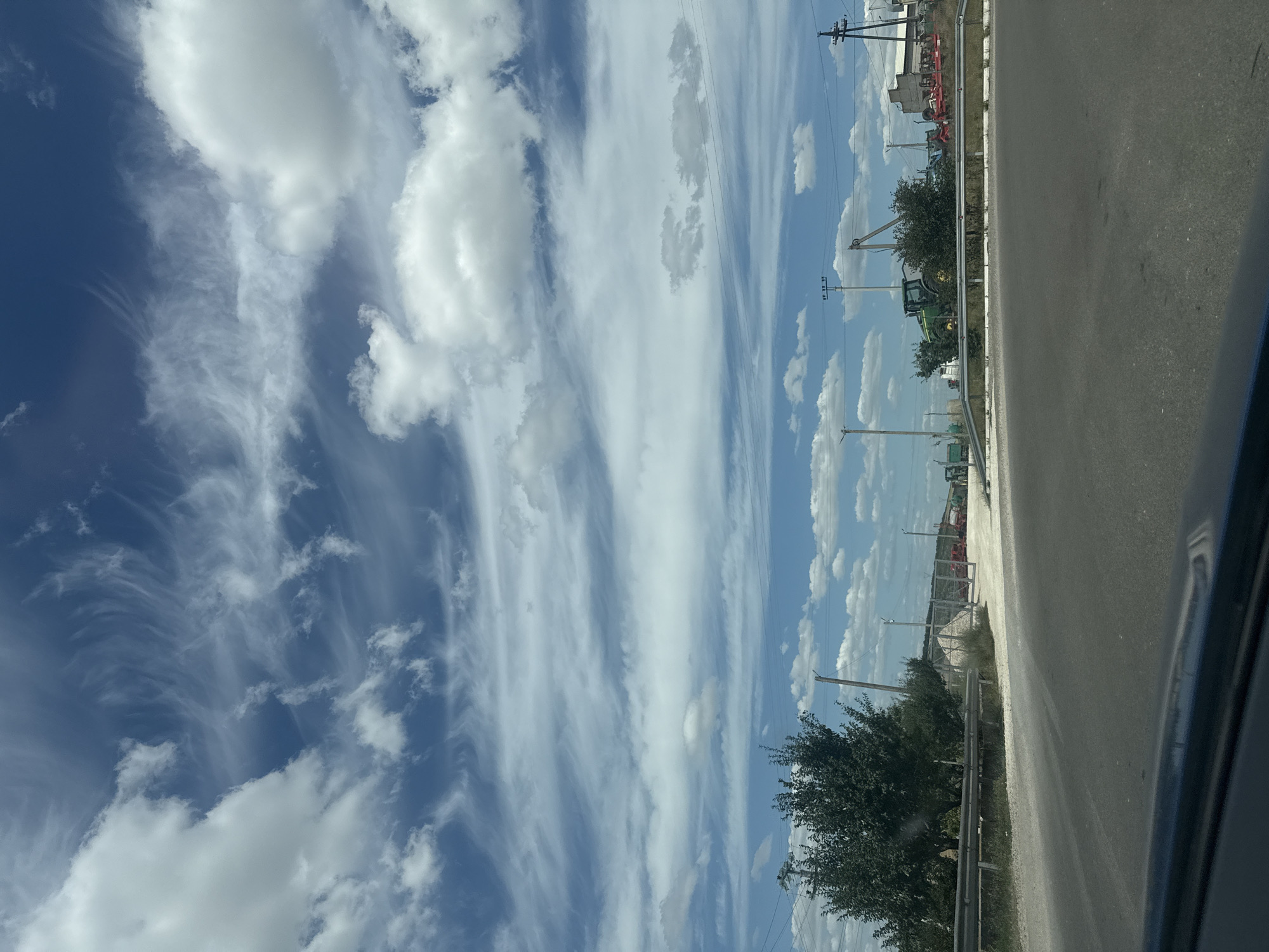 The image shows a roadside view in Iurievca, Moldova, on a cloudy day with a bright blue sky. The foreground features a section of asphalt road with a guardrail. Beyond the guardrail, there is a patch of dry grass and dirt. In the middle ground, there is an entrance to what appears to be an agricultural or industrial area, indicated by a fence and several pieces of heavy machinery. Visible equipment includes a green tractor with a loader, a red implement that looks like a harrow or plow, and other indistinct agricultural vehicles. The background is a mix of trees, power lines, and buildings. The sky is filled with a variety of clouds, some puffy and white, others thin and wispy, streaked across the blue.