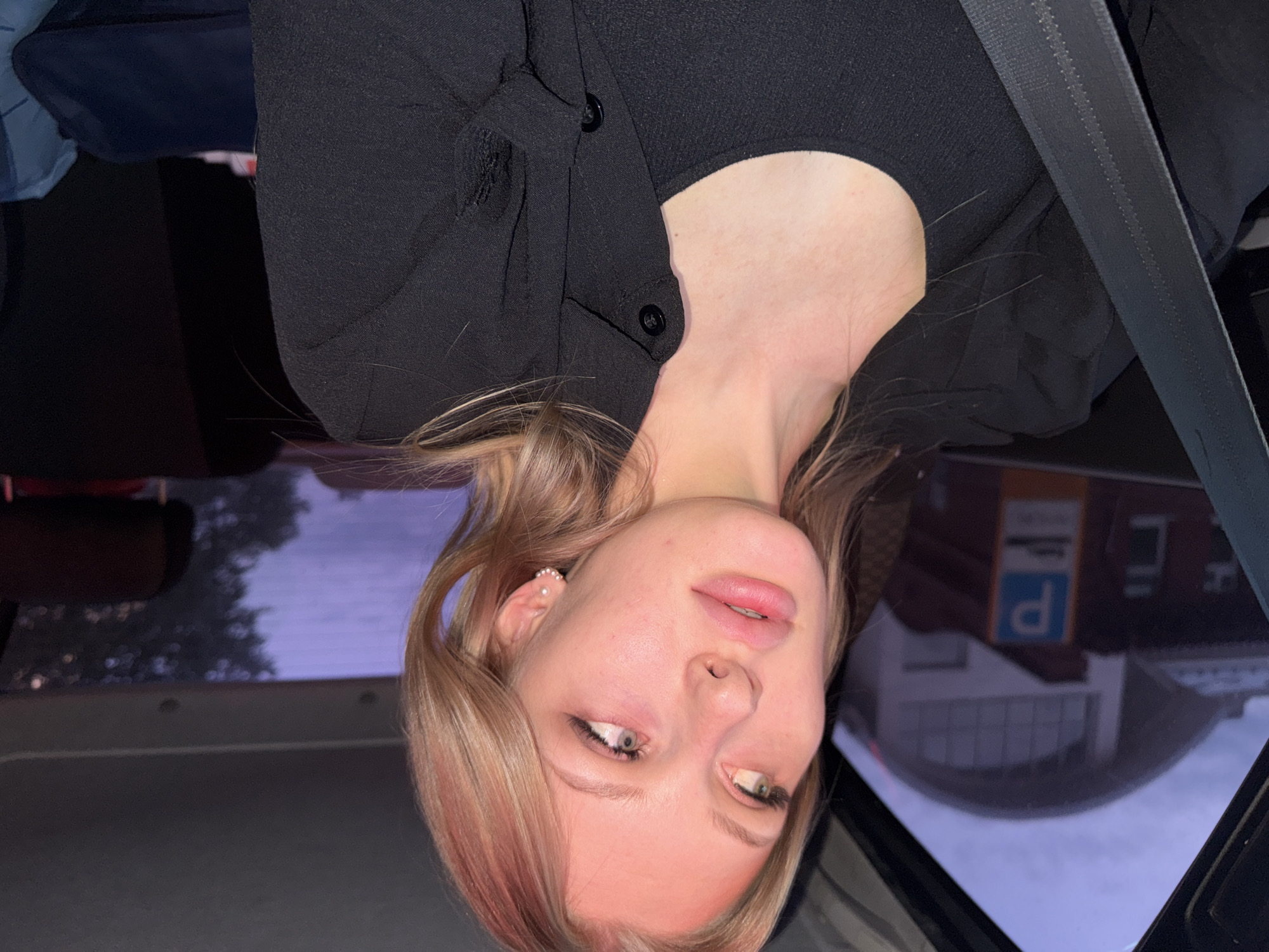 The image depicts a woman wearing a black shirt and a black jacket, likely in the passenger seat of a car, with a seatbelt across her torso. The image appears to be inverted. Through the car window, there is a reflection of a building in Perm, Russia, visible in the window, including a parking sign with the Cyrillic letter "P" displayed. The background shows a cloudy sky.