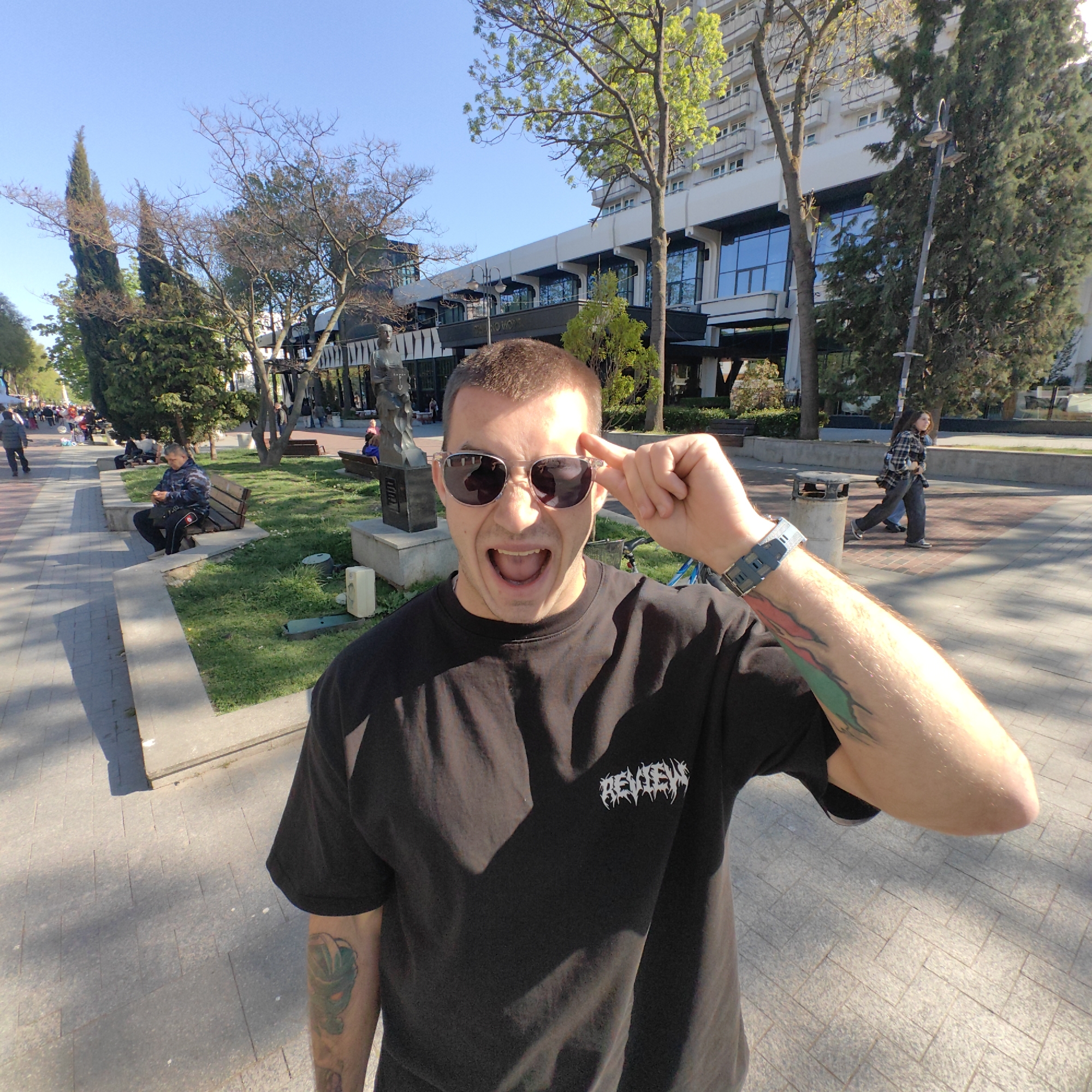 A man with tattoos and sunglasses stands in a public park. He wears a black t-shirt with white writing on the chest. He has his mouth open, with one hand raised to his head. The park has brick sidewalks, trees, benches, and statues. In the background is a multi-story white building with blue windows and balconies. Other people are walking around or sitting on benches. The park appears to be in Varna, Bulgaria, based on architectural style.