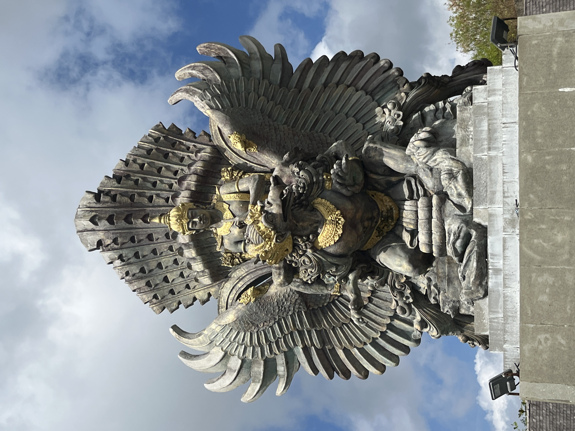 The image shows a close-up, low-angle view of the Garuda Wisnu Kencana statue in Kuta Selatan, Indonesia. The statue is monumental, made of stone with gold accents, and depicts a figure with large, feathered wings, a crown, and intricate jewelry. The figure is seated, with one hand gripping a smaller mythical creature or demon. The statue is positioned against a partially cloudy blue sky. The base of the statue is visible, with a multi-tiered stone platform. Two outdoor floodlights are mounted on the side of the platform. Trees are visible in the background on the right side of the image. The overall impression is one of grandeur and religious significance.